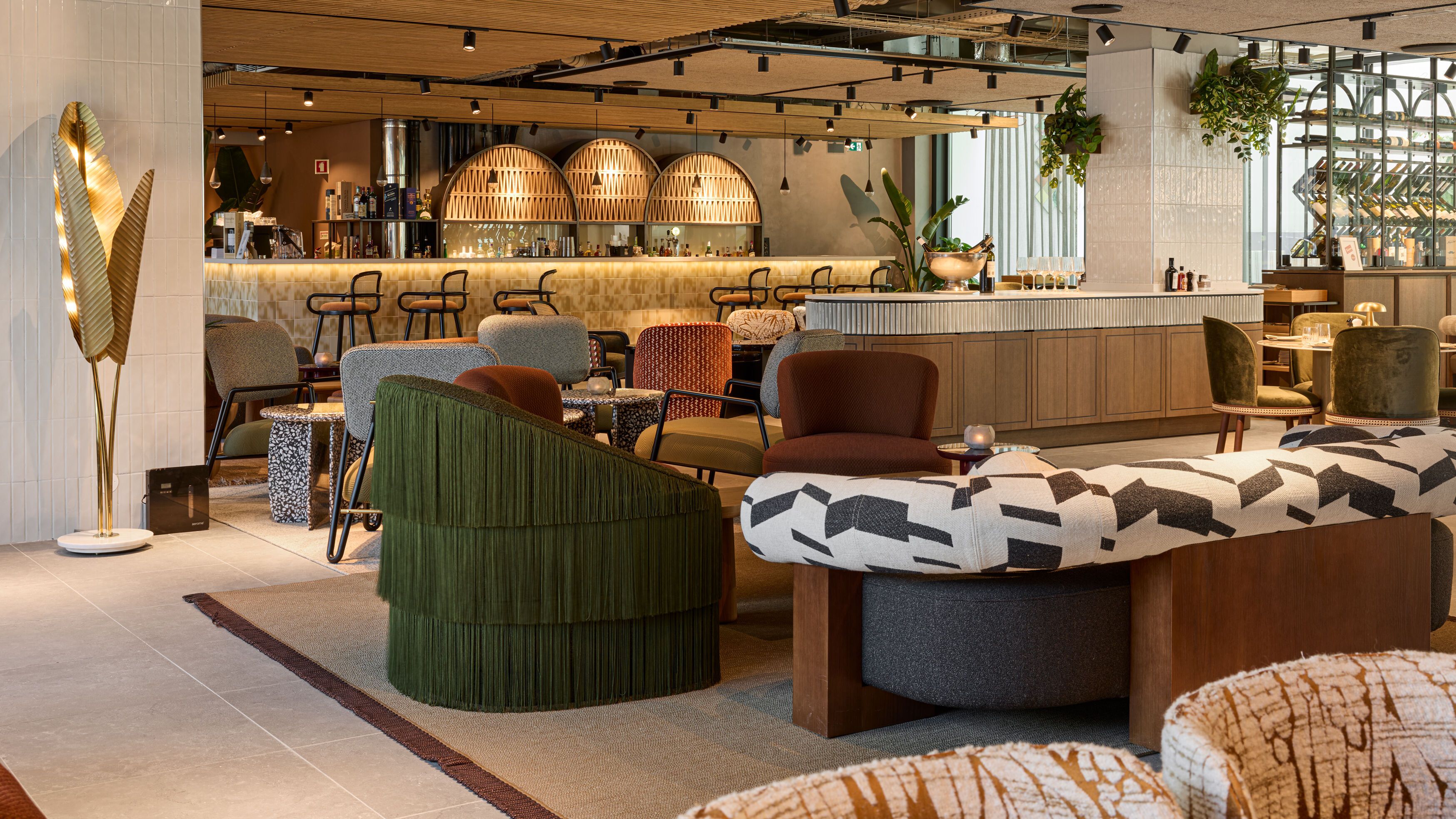 Moderne Hotelbar mit stilvollen Sitzgelegenheiten, warmem Holzdekor und atmosphärischer Beleuchtung. Gemütliche Lounge-Möbel laden zu entspannten Gesprächen bei einem Getränk ein, umgeben von grünen Pflanzen und elegantem Interieur.
