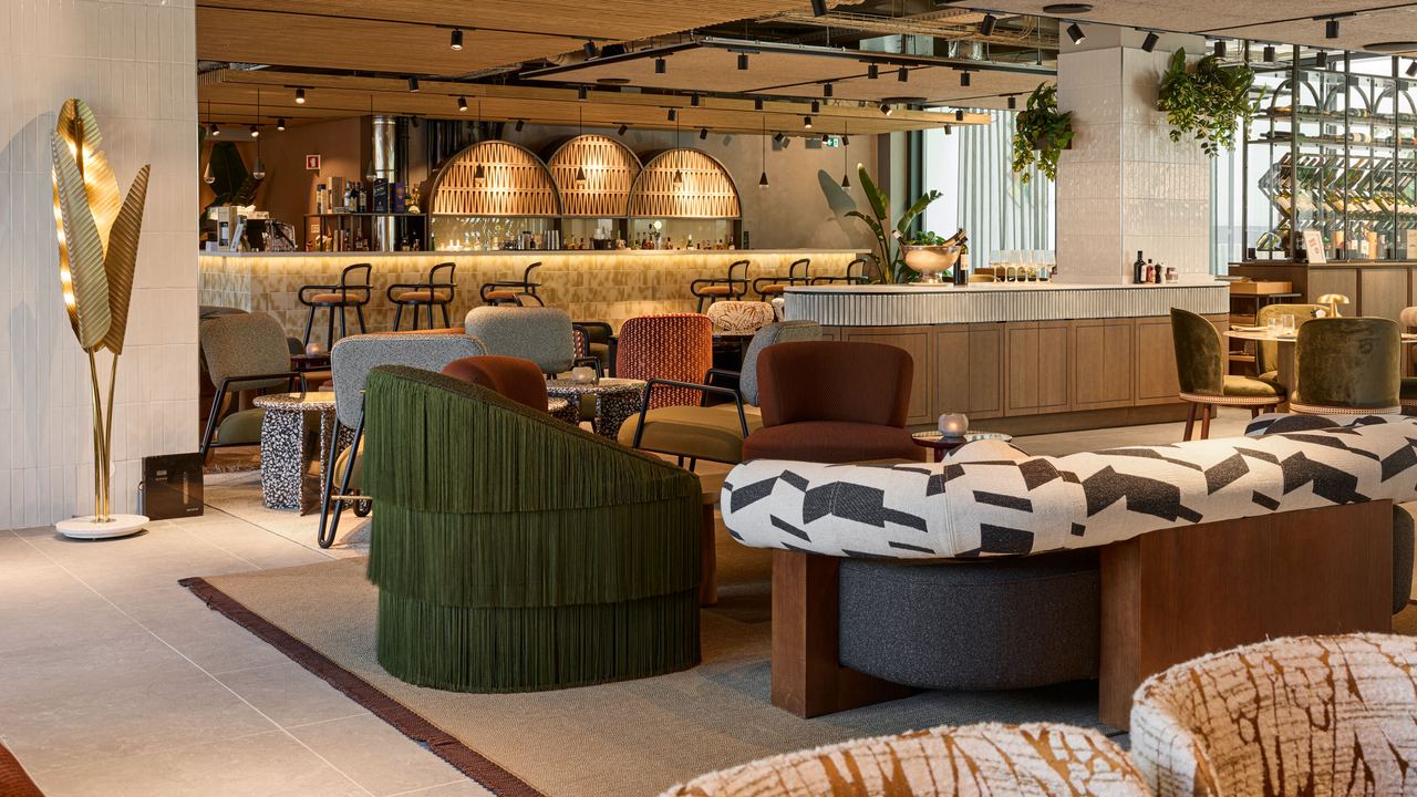 Moderne Hotelbar mit stilvollen Sitzgelegenheiten, warmem Holzdekor und atmosphärischer Beleuchtung. Gemütliche Lounge-Möbel laden zu entspannten Gesprächen bei einem Getränk ein, umgeben von grünen Pflanzen und elegantem Interieur.