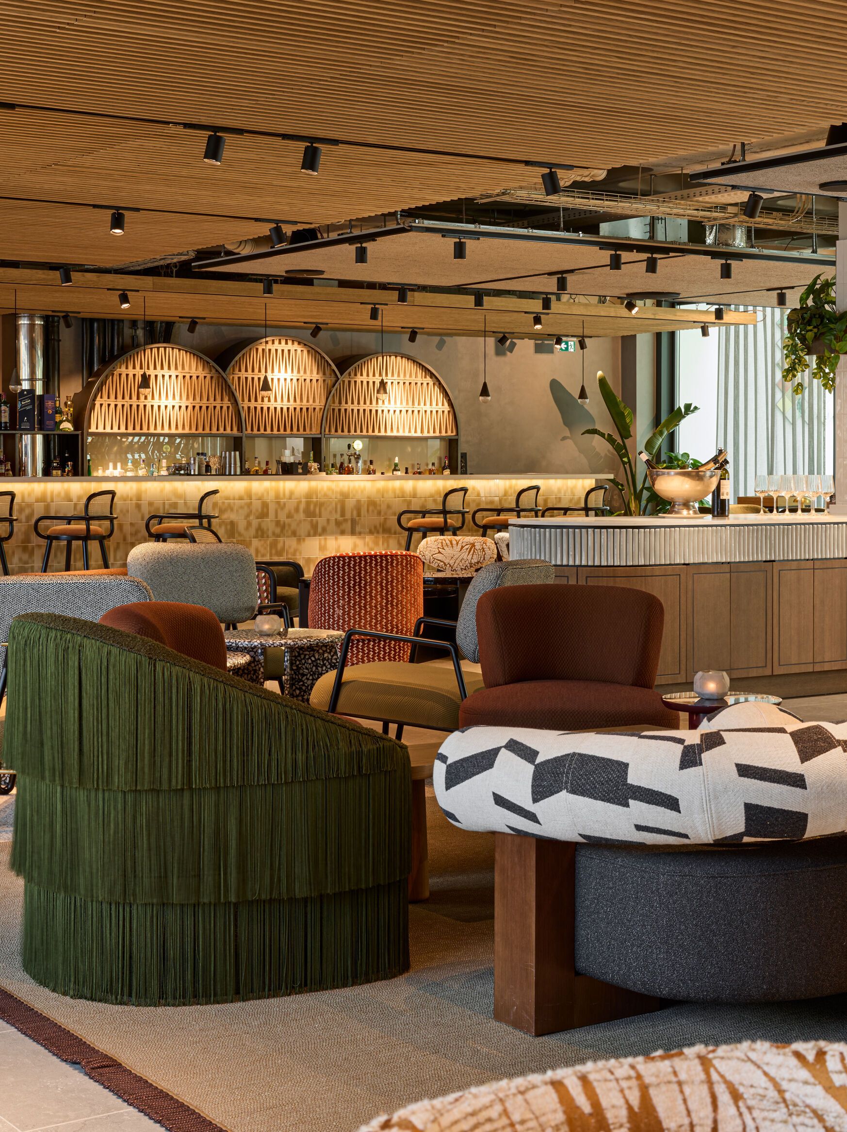 Moderne Hotelbar mit stilvollen Sitzgelegenheiten, warmem Holzdekor und atmosphärischer Beleuchtung. Gemütliche Lounge-Möbel laden zu entspannten Gesprächen bei einem Getränk ein, umgeben von grünen Pflanzen und elegantem Interieur.