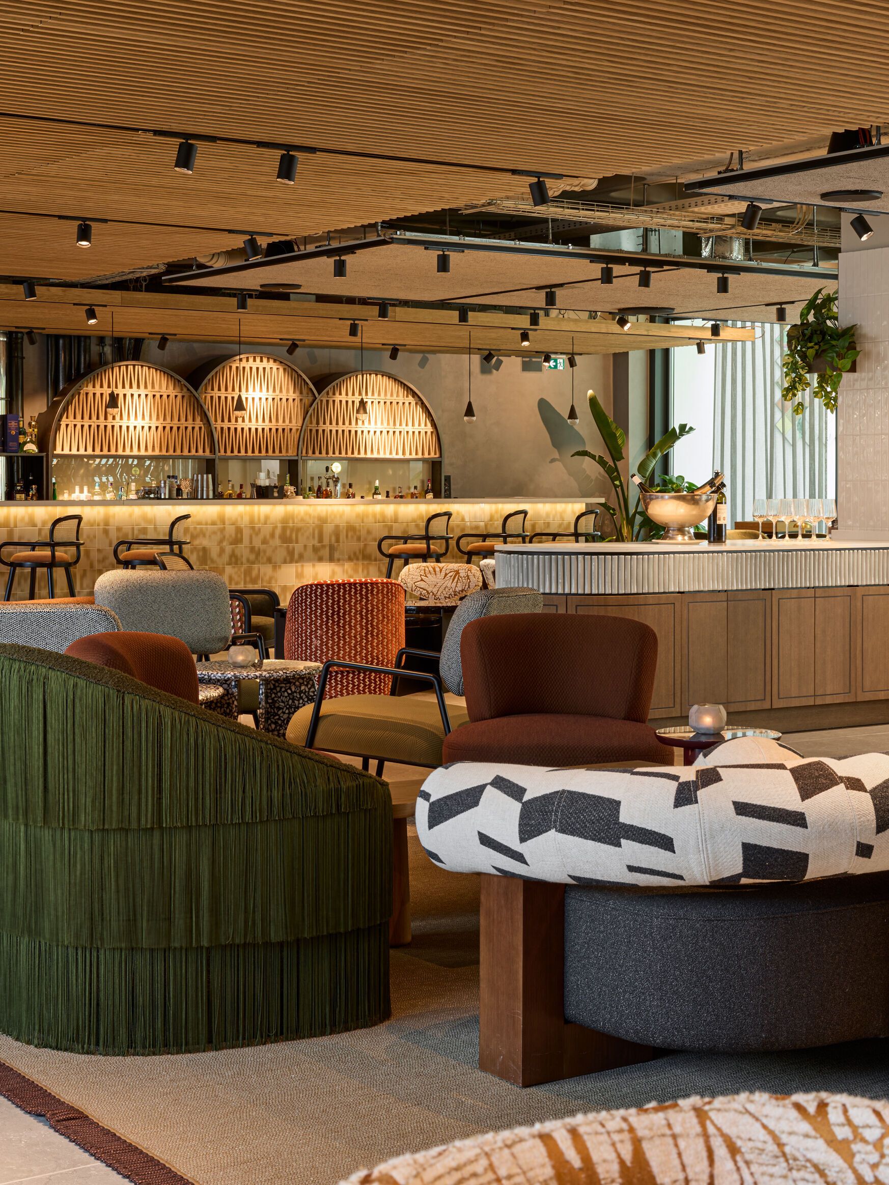 Moderne Hotelbar mit stilvollen Sitzgelegenheiten, warmem Holzdekor und atmosphärischer Beleuchtung. Gemütliche Lounge-Möbel laden zu entspannten Gesprächen bei einem Getränk ein, umgeben von grünen Pflanzen und elegantem Interieur.