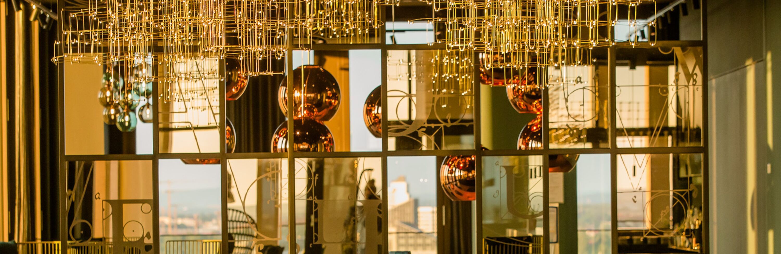 Blick durch ein modernes Spiegelpaneel, das den Berliner Fernsehturm und andere Gebäude in warmem Abendlicht reflektiert. Hängeleuchten mit goldfarbenen Röhren
