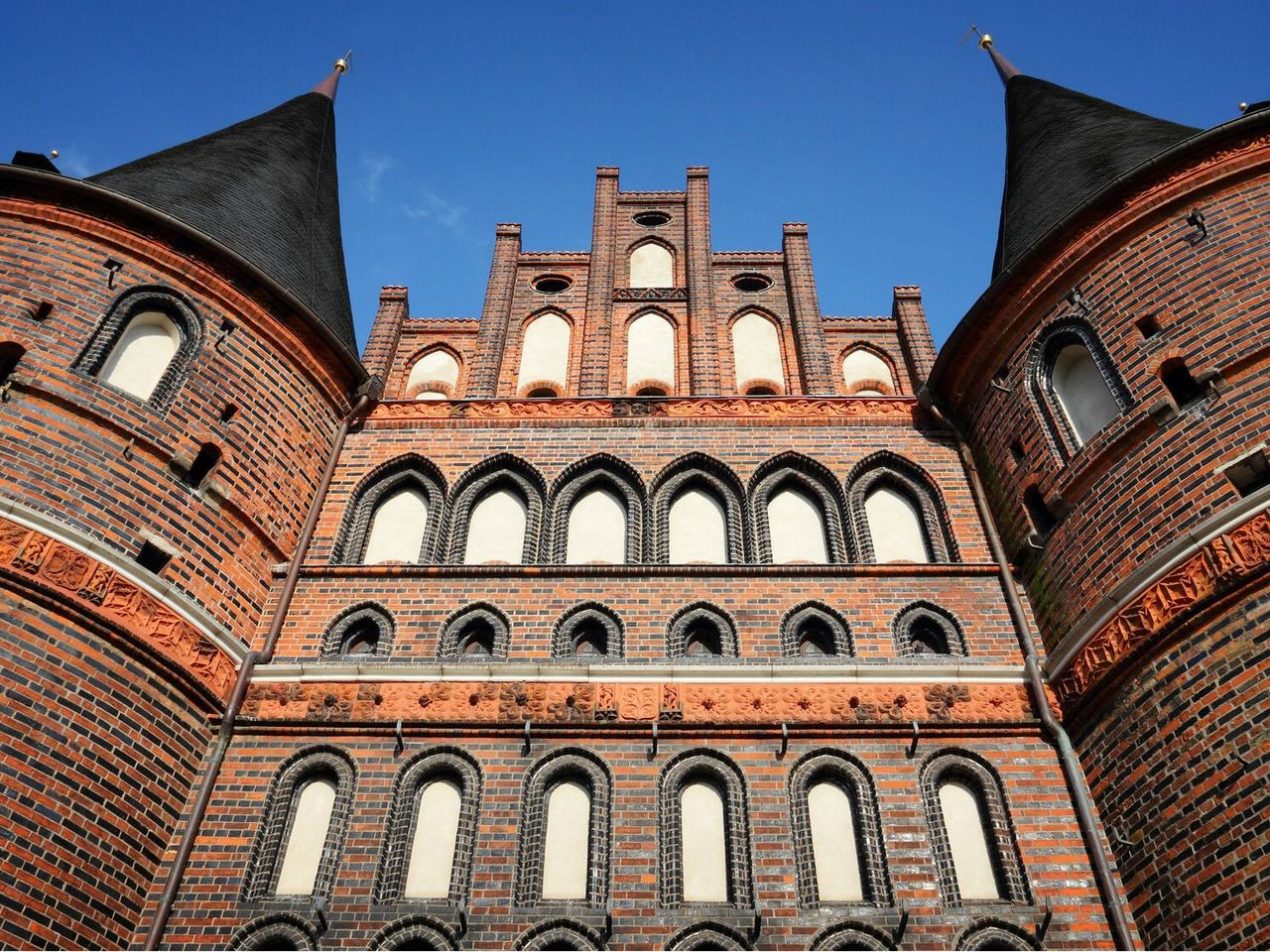 Backsteingotik-Fassade mit zwei runden Ecktürmen und spitzen Dächern, zahlreichen hohen, schmalen Fenstern und dekorativen Ziegelornamenten unter strahlend blauem Himmel.