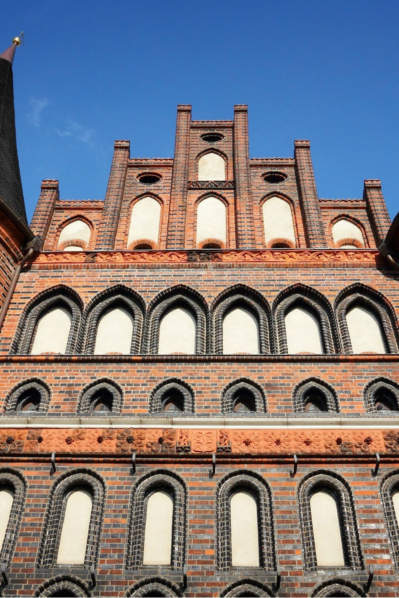 Backsteingotik-Fassade mit zwei runden Ecktürmen und spitzen Dächern, zahlreichen hohen, schmalen Fenstern und dekorativen Ziegelornamenten unter strahlend blauem Himmel.
