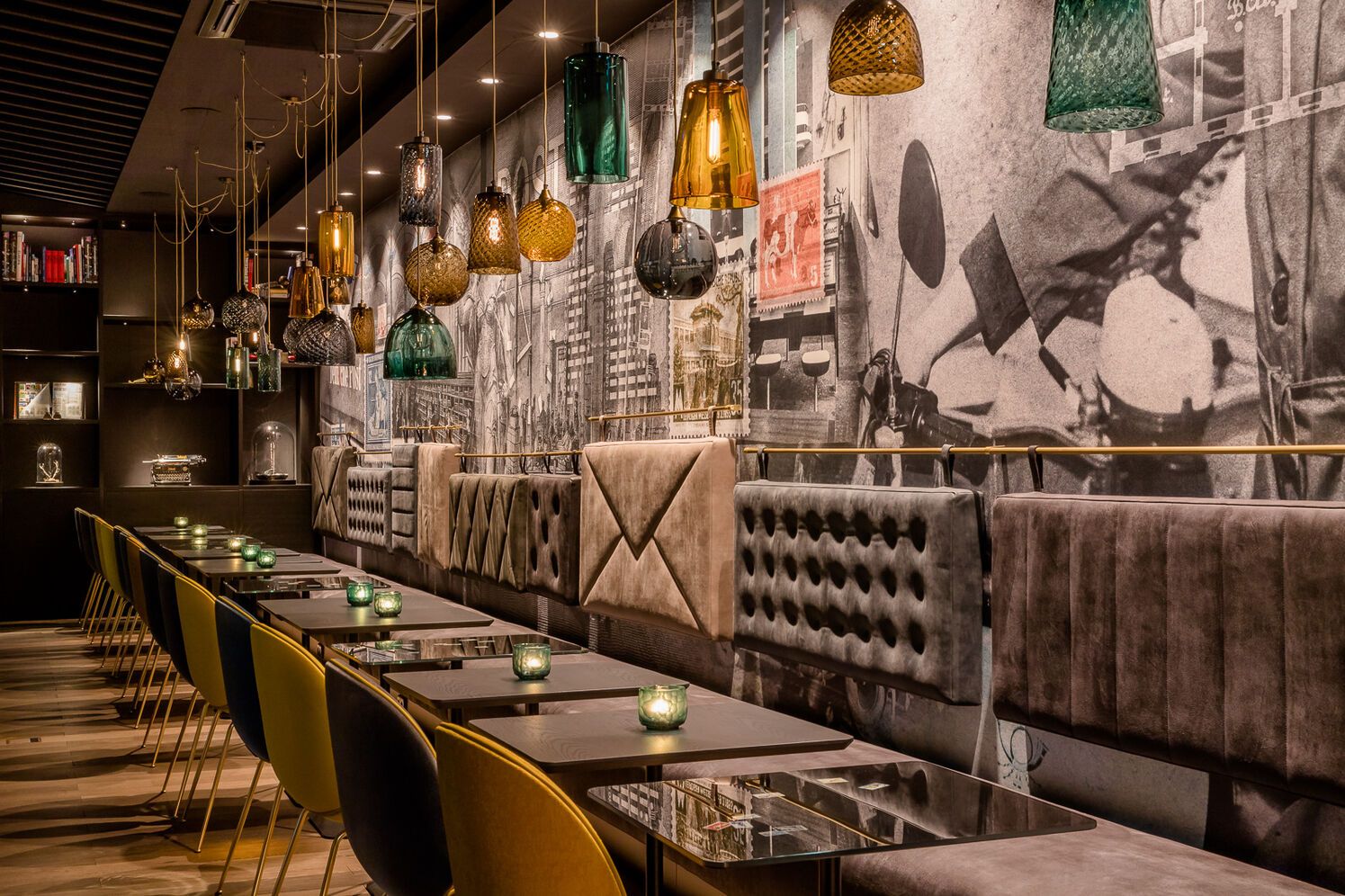 Gemütlicher Hotel-Barbereich mit samtbezogenen Sitzbänken, eleganten Stühlen und warm leuchtenden Hängelampen vor einer großformatigen, urban inspirierten Wandgestaltung.