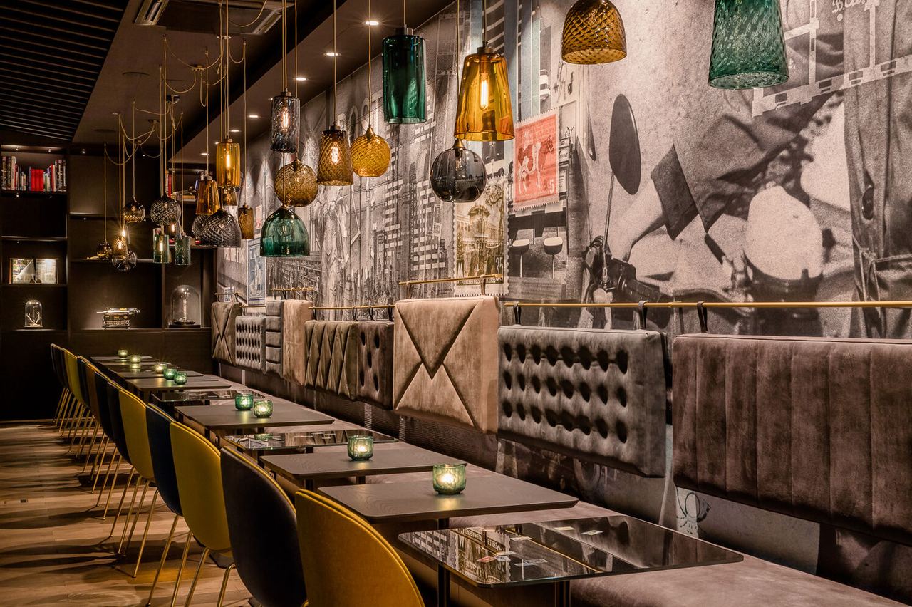 Gemütlicher Hotel-Barbereich mit samtbezogenen Sitzbänken, eleganten Stühlen und warm leuchtenden Hängelampen vor einer großformatigen, urban inspirierten Wandgestaltung.