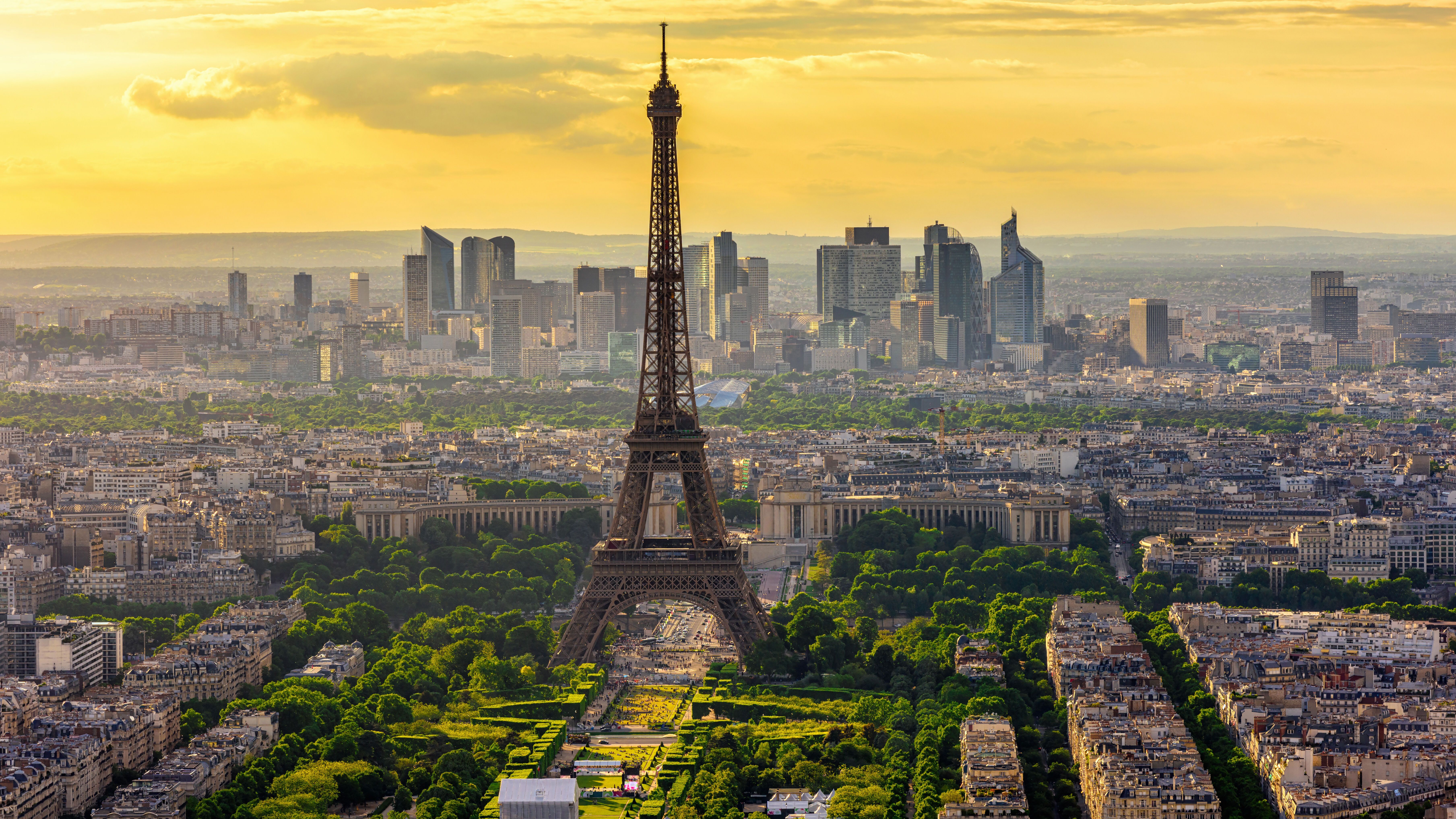 Panoramablick auf den Eiffelturm in Paris bei Sonnenuntergang, umgeben von grünen Parks und der modernen Skyline von La Défense im Hintergrund.