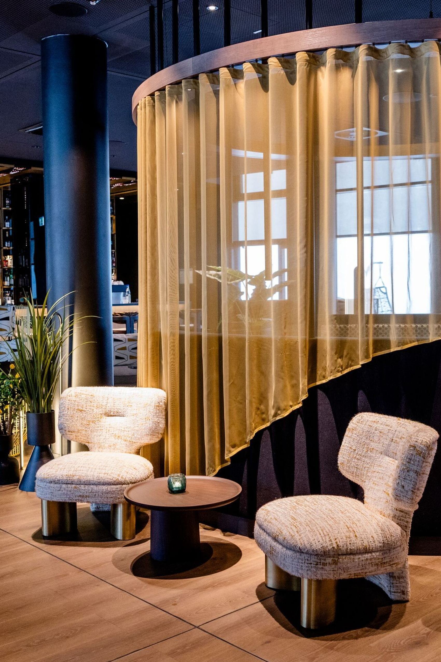 Hotel Frankfurt-Messe Motel One Lounge und Bar mit Sitzgruppe aus bequemen Sesseln, rundem Tisch und halbtransparentem Vorhang. Warme Beleuchtung und elegante Holz- und Stoffelemente.