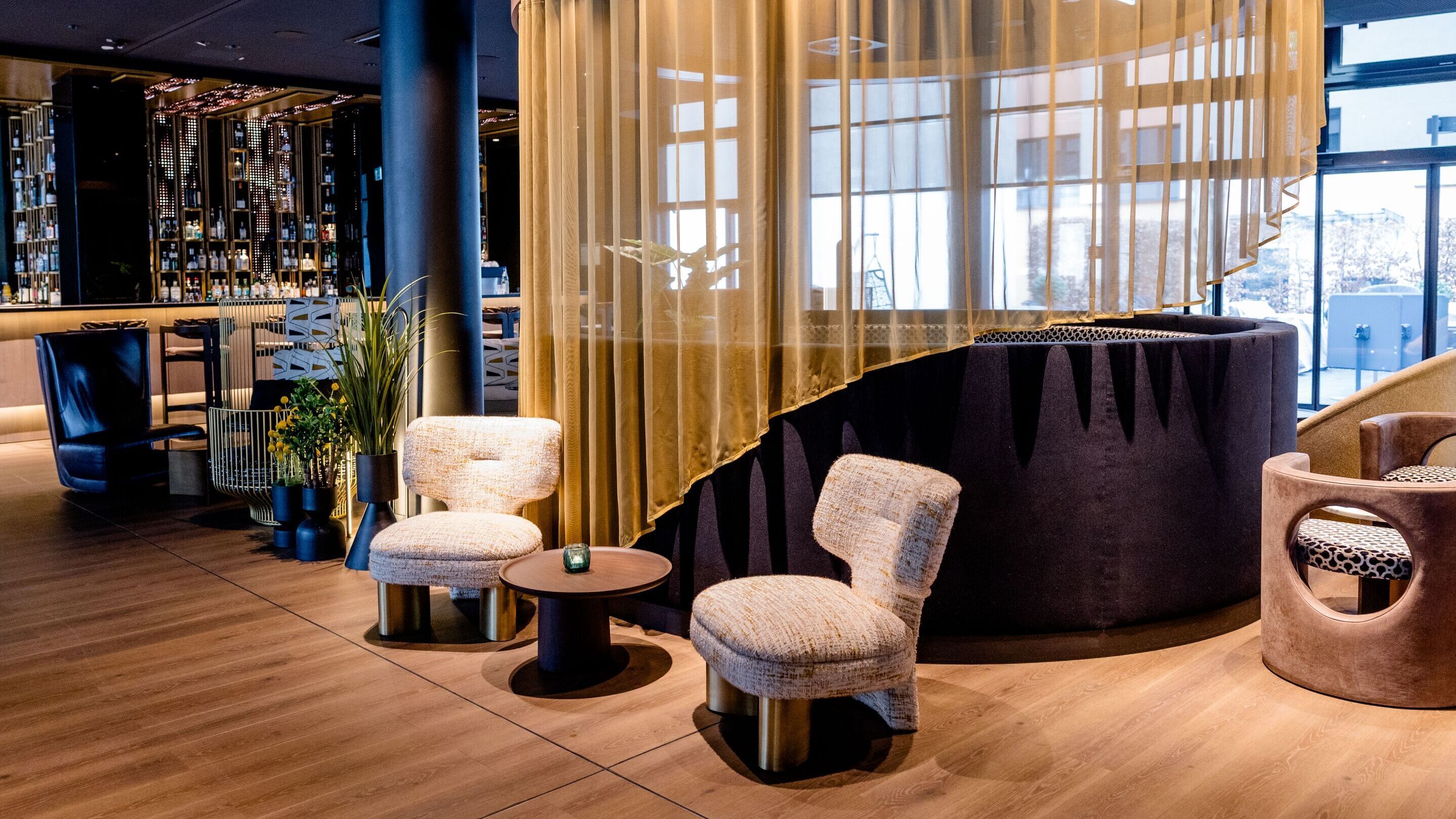 Hotel Frankfurt-Messe Motel One lounge and bar with seating area featuring comfortable armchairs, round table and semi-transparent curtain. Warm lighting and elegant wood and fabric elements.