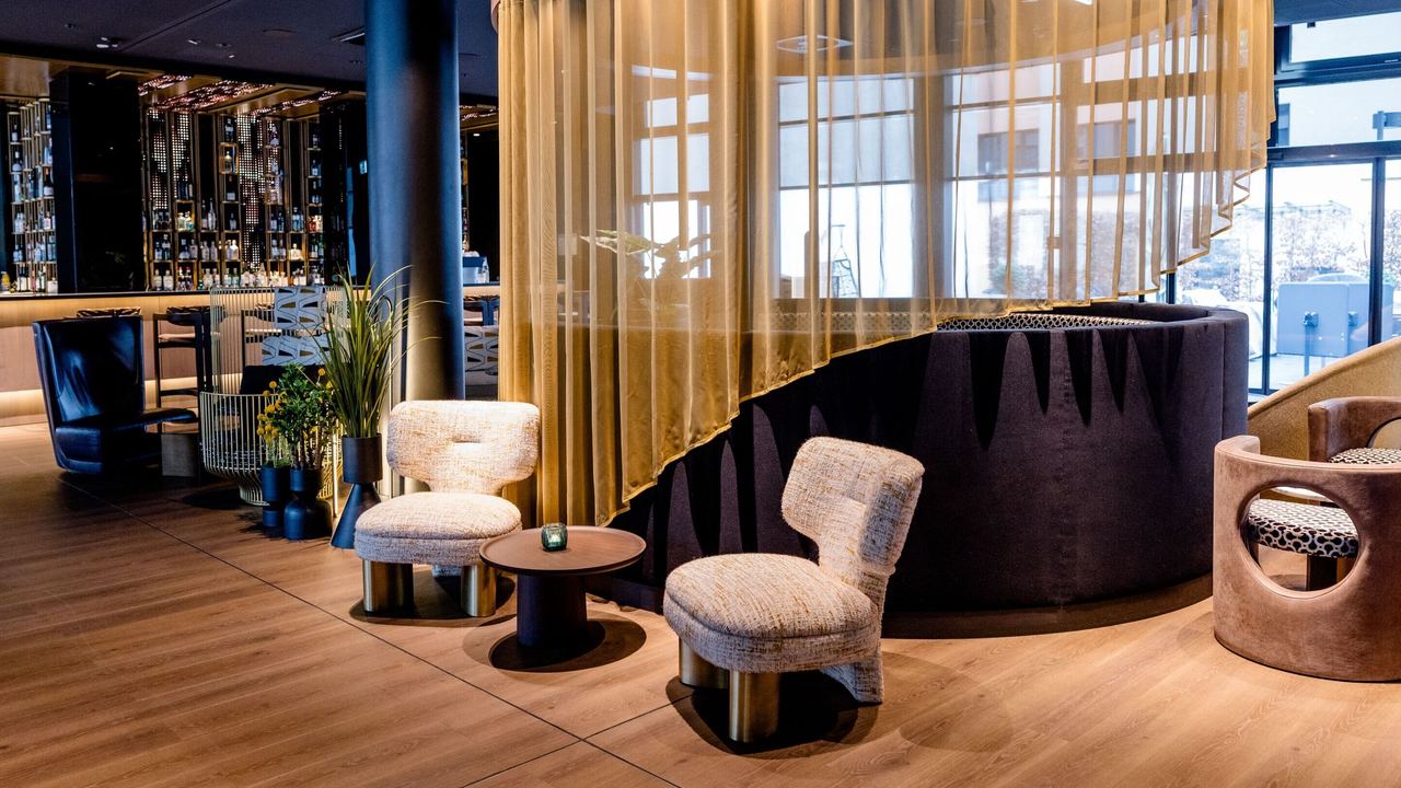 Hotel Frankfurt-Messe Motel One lounge and bar with seating area featuring comfortable armchairs, round table and semi-transparent curtain. Warm lighting and elegant wood and fabric elements.