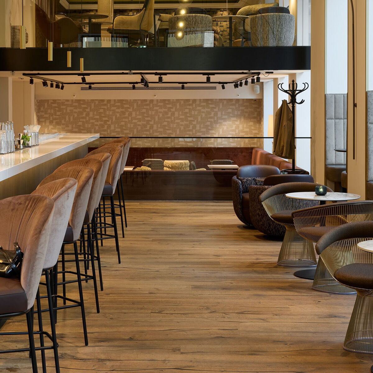 Moderner Hotelbarbereich mit langer Sitzreihe an der Theke, gemütlichen Lounge-Sesseln und einer offenen Empore mit zusätzlichen Sitzgelegenheiten, warmes Holzdesign und stilvolle Beleuchtung schaffen ein einladendes Ambiente für entspannte Stunden.