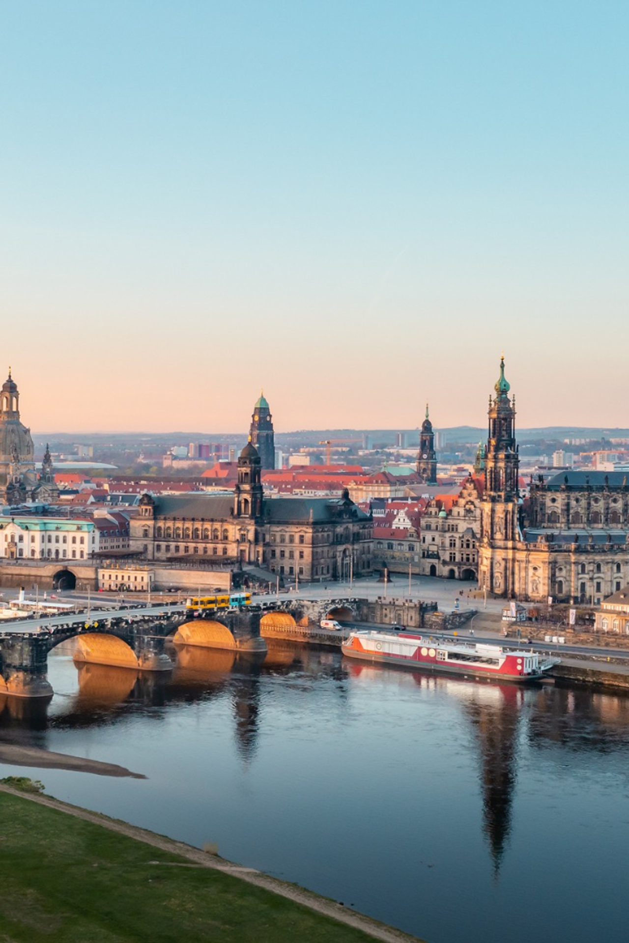 Panoramablick auf die historische Altstadt von Dresden mit der Elbe, einer Brücke und markanten barocken Türmen im sanften Licht des Sonnenuntergangs.
