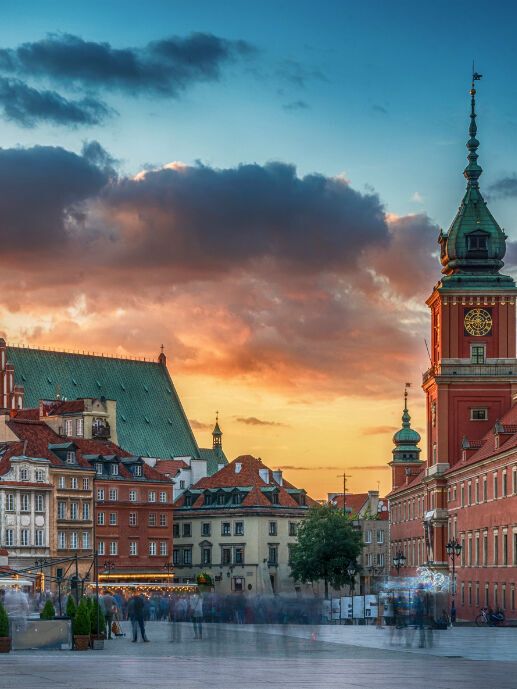 Historische Altstadt mit farbenfrohen Häusern und turmhohen Bauwerken im warmen Abendlicht, malerischer Sonnenuntergang am Himmel, lebendiger Marktplatz mit Menschen in Bewegung.