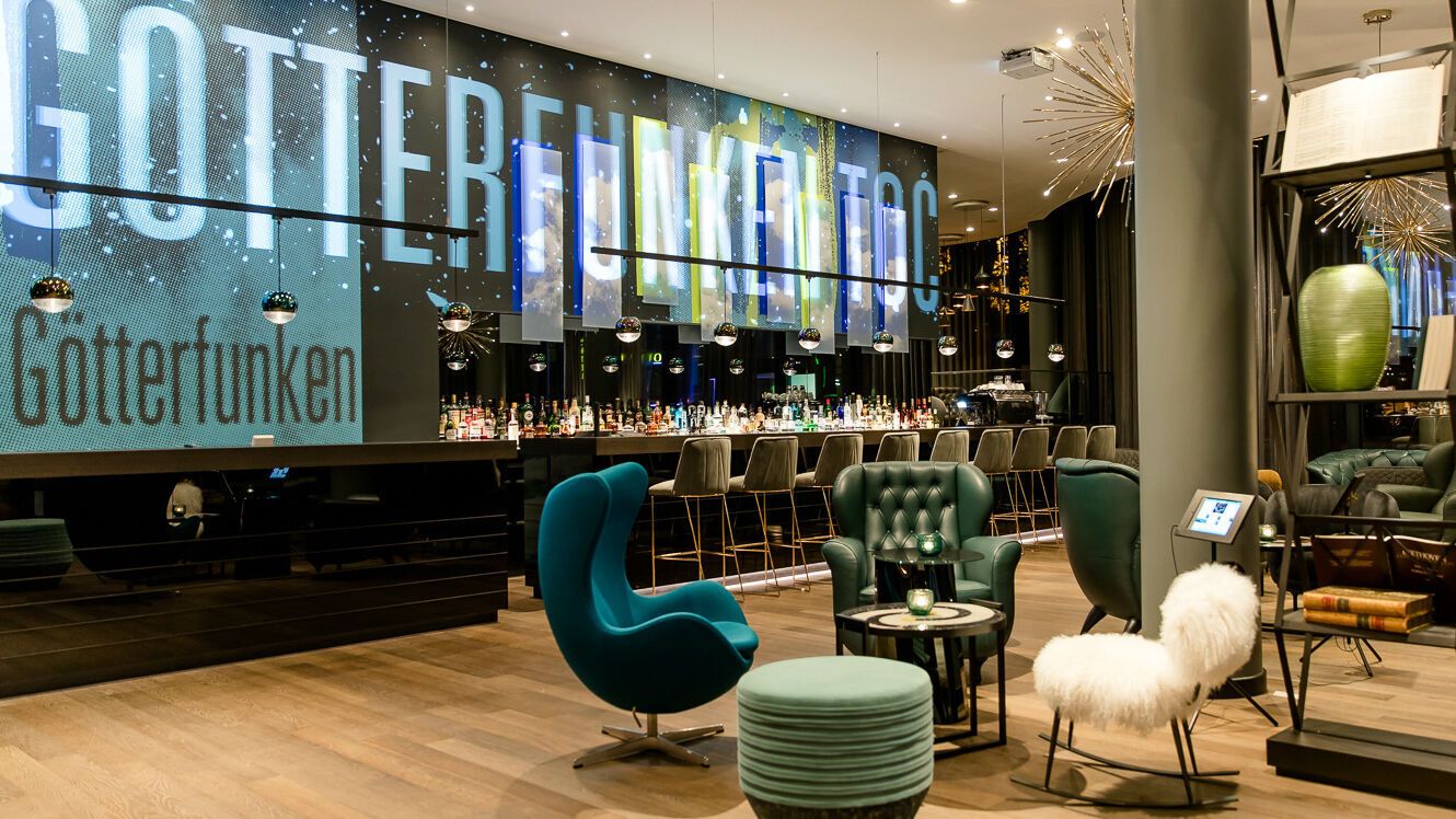Motel One Hotelbar mit stilvoller Beleuchtung, gemütlichen Sitzgelegenheiten und einer langen Theke mit Barhockern. Großes, beleuchtetes Schriftzug-Display an der Wand.