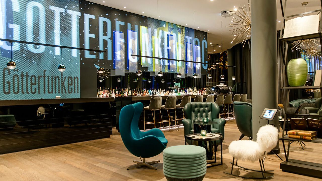 Motel One Hotelbar mit stilvoller Beleuchtung, gemütlichen Sitzgelegenheiten und einer langen Theke mit Barhockern. Großes, beleuchtetes Schriftzug-Display an der Wand.