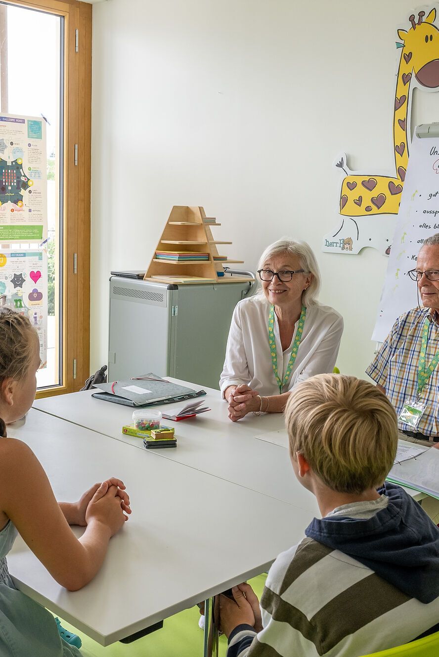 Sechs Kinder sitzen an einem Tisch in einem hellen Raum und hören gespannt zwei Erwachsenen zu, die eine Gruppe anleiten. Im Hintergrund ist eine Giraffen-Dekoration und ein Flipchart mit handgeschriebenen Notizen zu sehen.