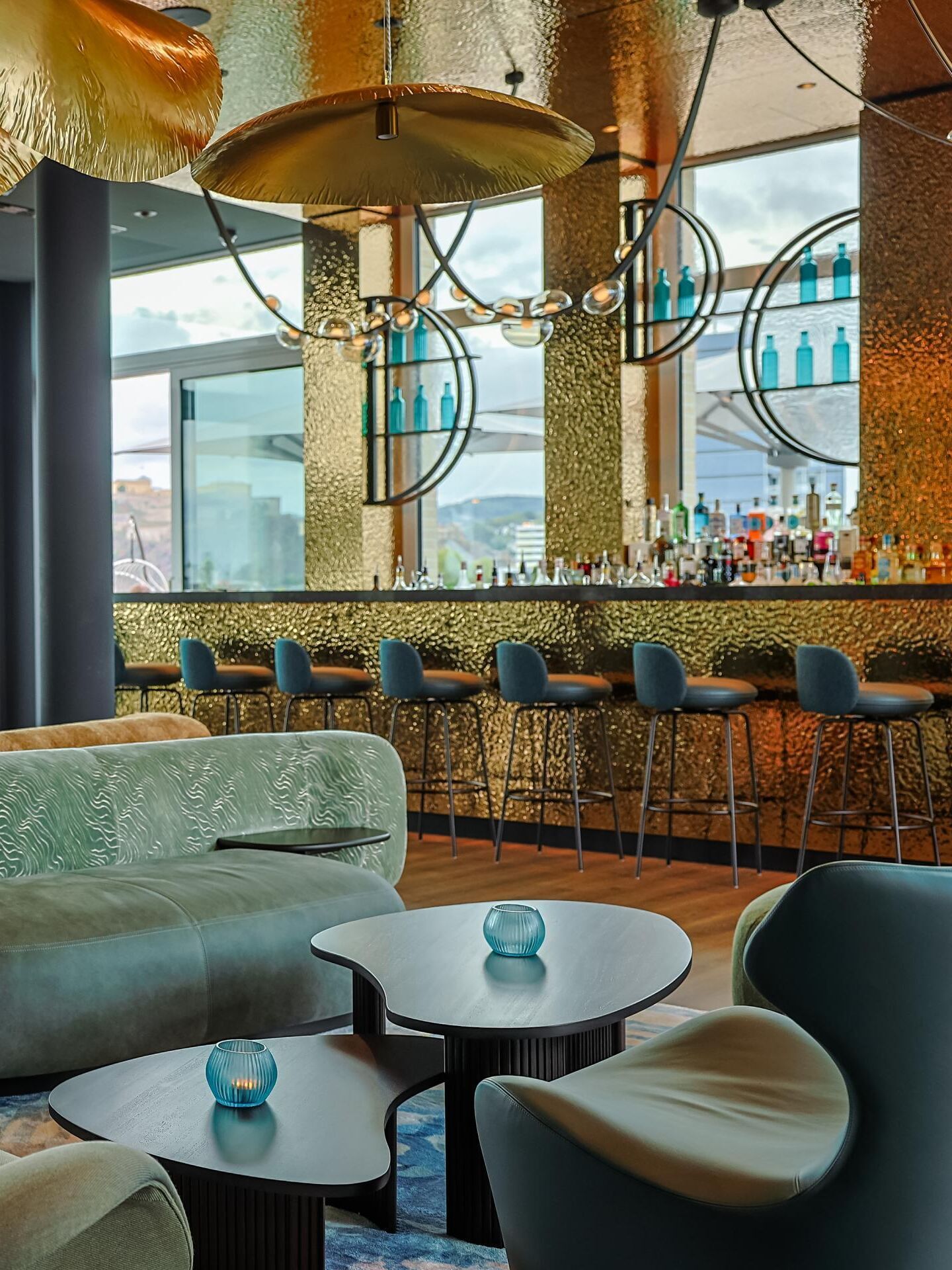 Modern gestaltete Hotelbar mit bequemen Sesseln und Sofas, großen Fenstern mit Stadtblick, auffälligen goldfarbenen Deckenleuchten und einer langen Bar mit Barhockern, die zum entspannten Verweilen einlädt.