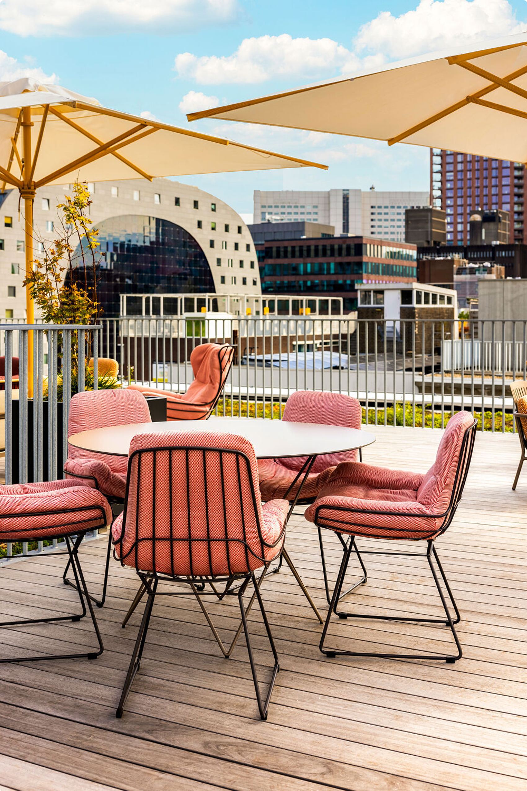 Dachterrasse mit gemütlichen, rosa gepolsterten Stühlen und rundem Tisch, großer Sonnenschirme und moderner Stadtkulisse im Hintergrund, perfekt zum Entspannen oder Essen im Freien.