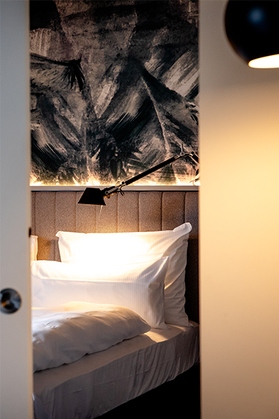 Blick durch geöffnete Zimmertür auf ein einladendes Bett mit weißen Kissen und Decke, sanfte Beleuchtung und moderne, künstlerische Wandgestaltung im Hintergrund.