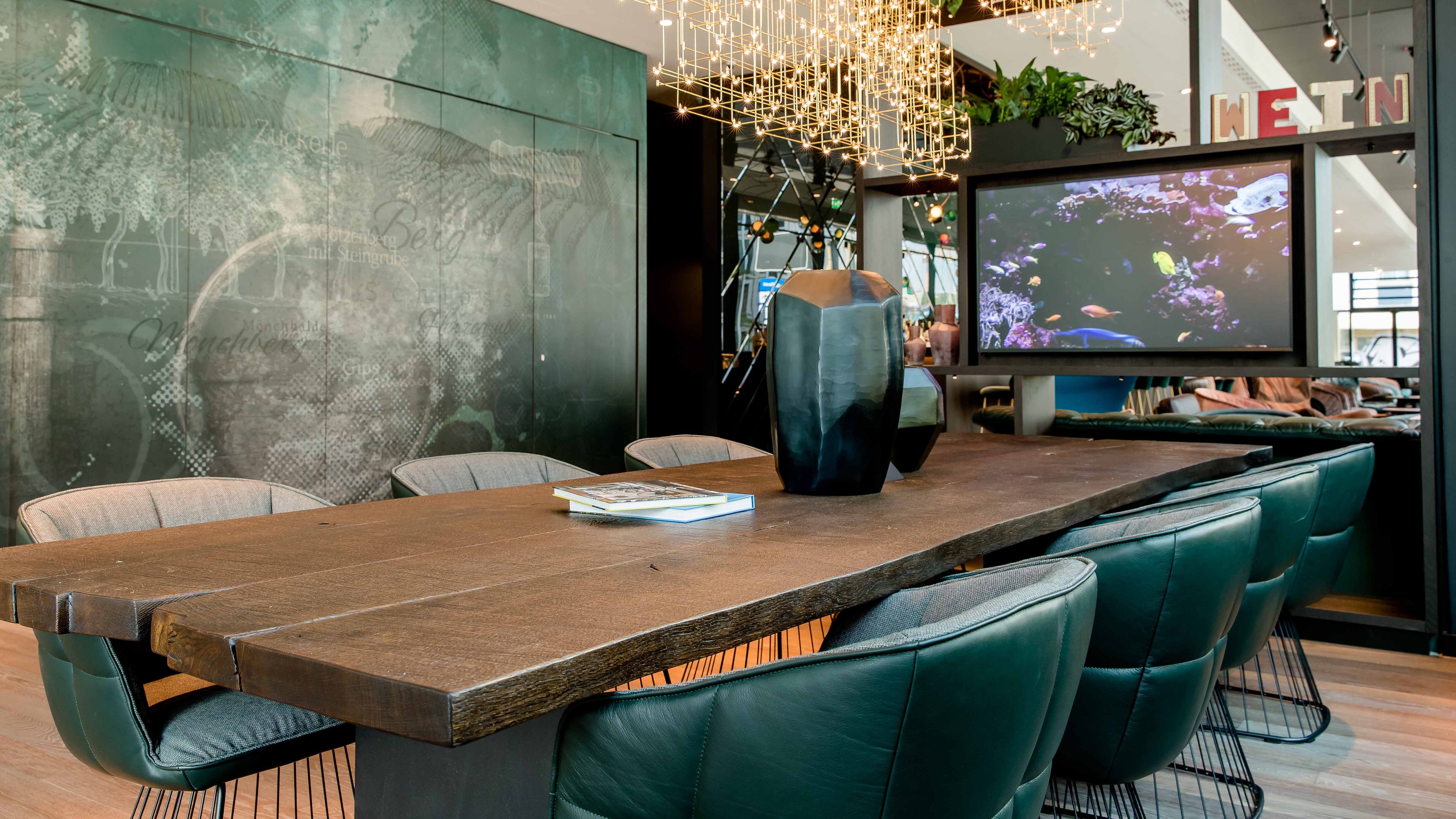 Motel One Konferenzraum mit langem Holztisch, bequemen Lederstühlen, stilvollem Blumendekor und indirekter Beleuchtung, ideal für Meetings oder entspannte Gespräche in einem eleganten Ambiente.