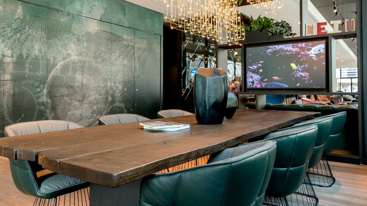 Motel One Konferenzraum mit langem Holztisch, bequemen Lederstühlen, stilvollem Blumendekor und indirekter Beleuchtung, ideal für Meetings oder entspannte Gespräche in einem eleganten Ambiente.