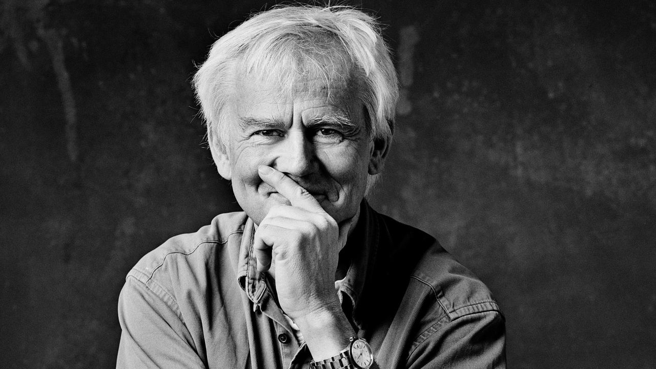 Porträt eines älteren Mannes mit hellem Haar, der nachdenklich lächelt und die rechte Hand leicht vor den Mund hält.