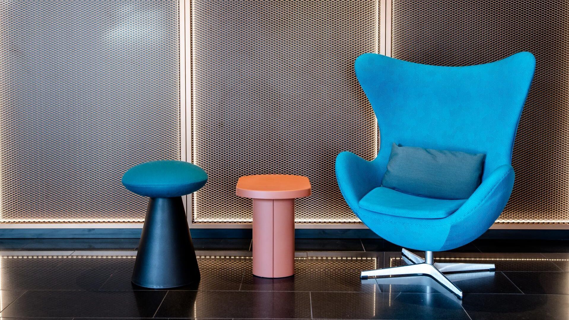 Moderner, gemütlicher Sitzbereich mit einem auffällig blauen Sessel und zwei kleinen Beistelltischen vor einer beleuchteten, metallischen Wand, ideal zum Entspannen oder für kurze Gespräche.
