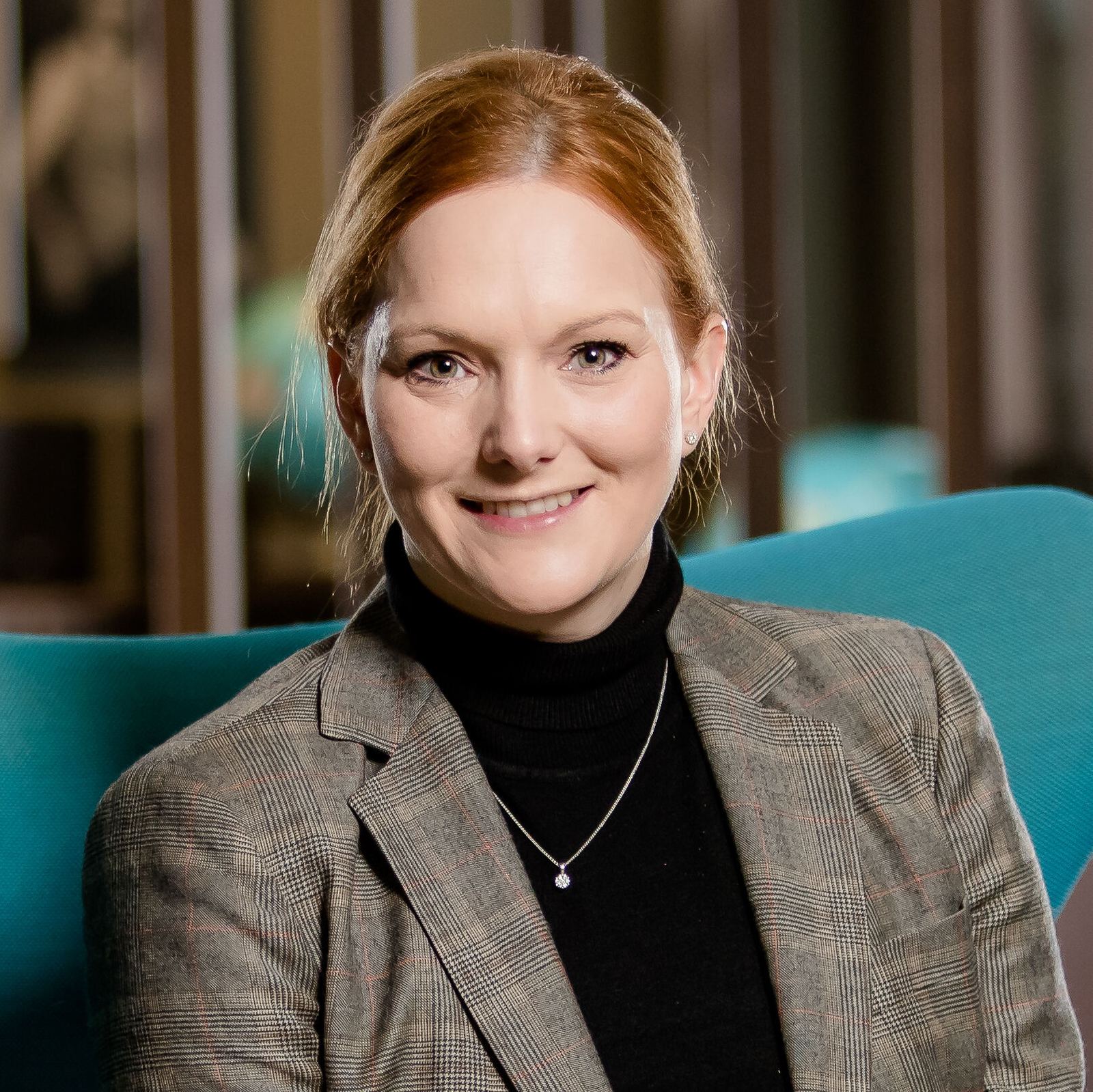 Eine freundlich lächelnde Frau in eleganter Kleidung sitzt entspannt in einem modernen, blauen Sessel in einer stilvollen Hotellobby mit warmem Ambiente.