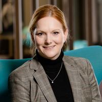 Eine freundlich lächelnde Frau in eleganter Kleidung sitzt entspannt in einem modernen, blauen Sessel in einer stilvollen Hotellobby mit warmem Ambiente.