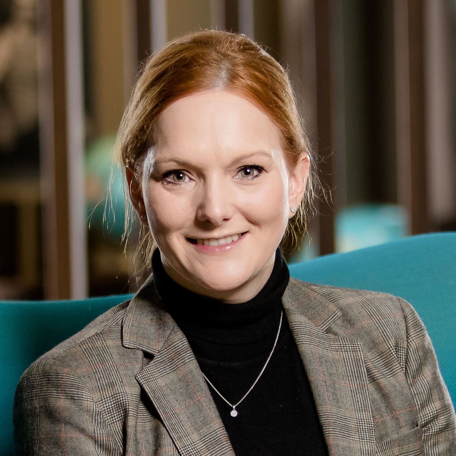 Eine freundlich lächelnde Frau in eleganter Kleidung sitzt entspannt in einem modernen, blauen Sessel in einer stilvollen Hotellobby mit warmem Ambiente.