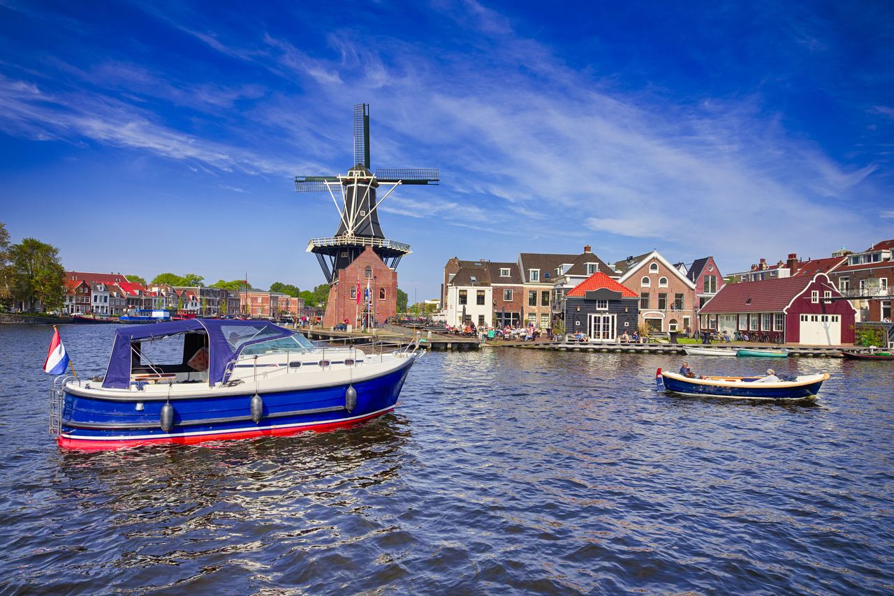 Traditionelle holländische Windmühle am Ufer eines ruhigen Kanals, umgeben von historischen Häusern und zwei Booten auf dem Wasser, unter einem klaren blauen Himmel.