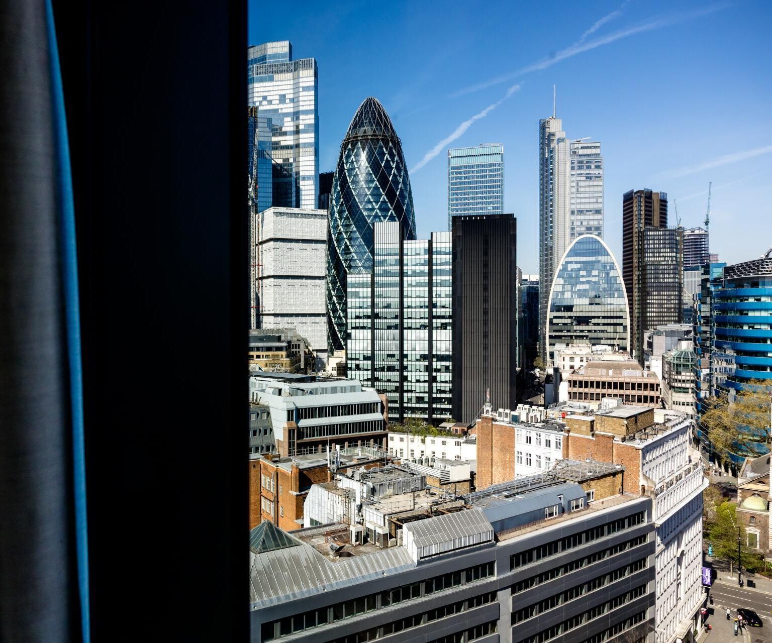 Blick aus einem Hotelfenster auf die moderne Skyline Londons mit markanten Hochhäusern wie dem Gherkin, bei klarem Himmel.