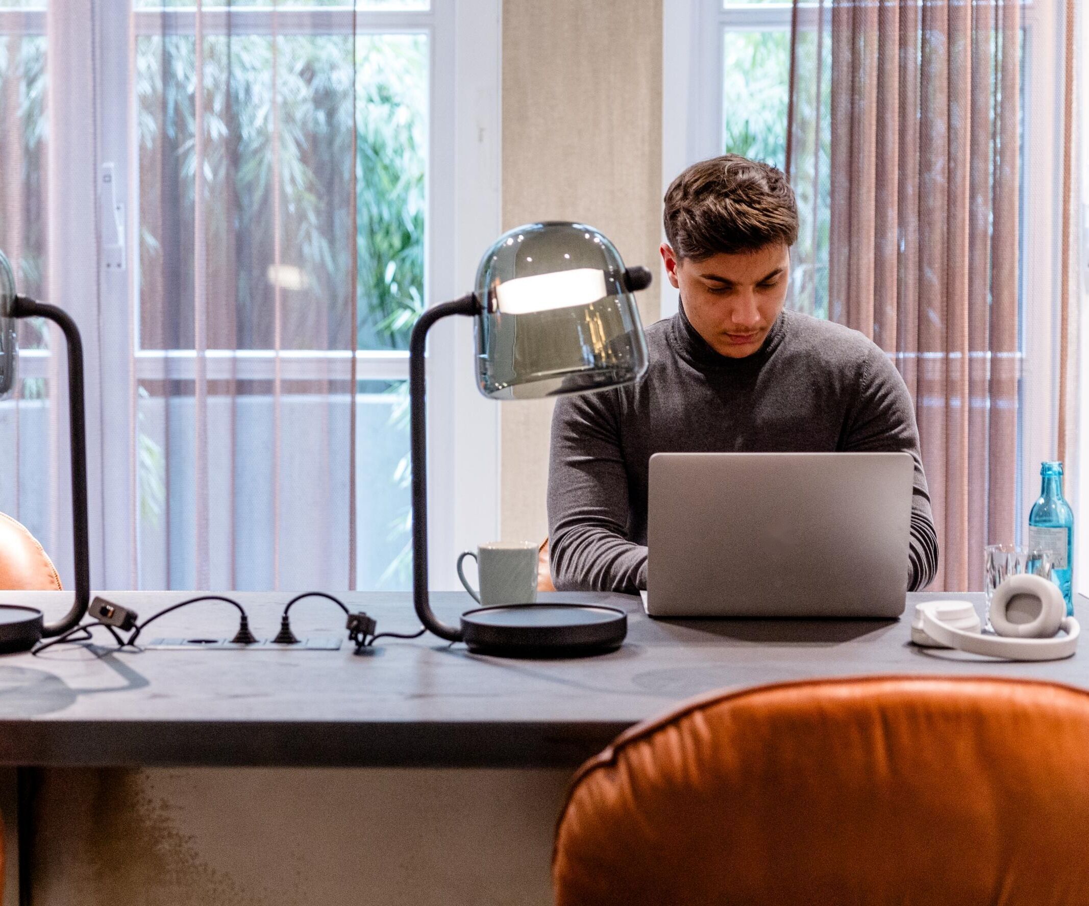Junger Mann arbeitet konzentriert an Laptop an einem modernen Arbeitstisch mit Lederstühlen und Schreibtischlampen vor großen Fenstern.
