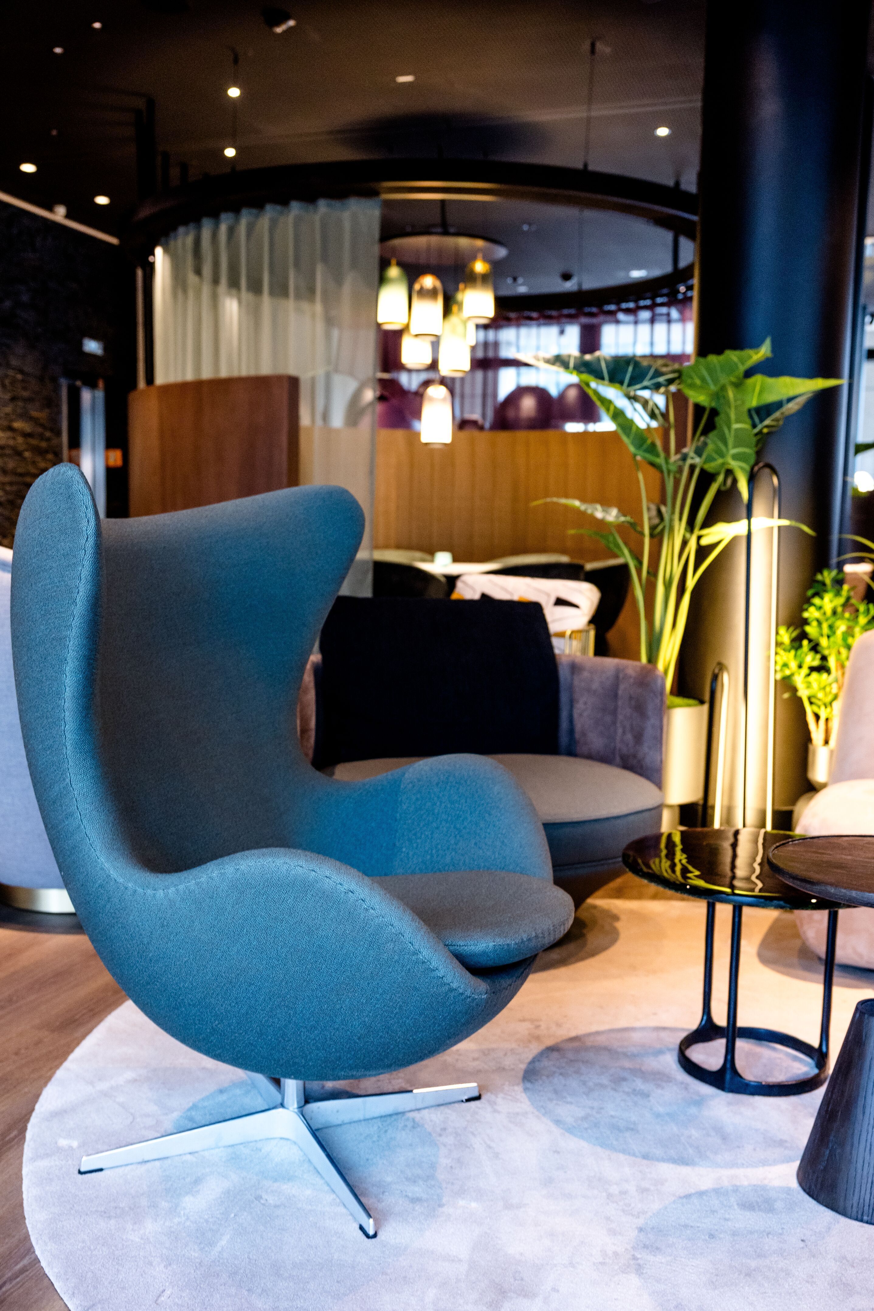 Hotel Frankfurt-Messe Motel One Lounge with Egg Chair in the foreground, round rug, elegant side tables, soothing lighting and green plants.