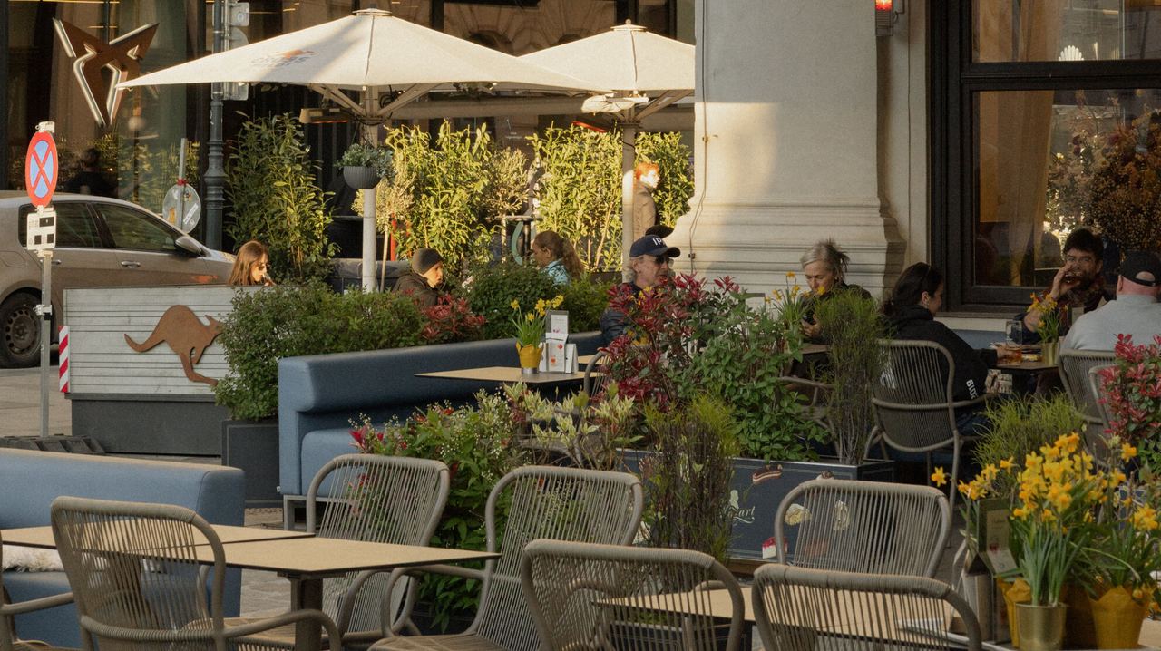 Außenbereich eines Cafés mit Sonnenschirmen und Sitzgelegenheiten, Menschen genießen bei warmem Licht Speisen und Getränke in entspannter Atmosphäre.