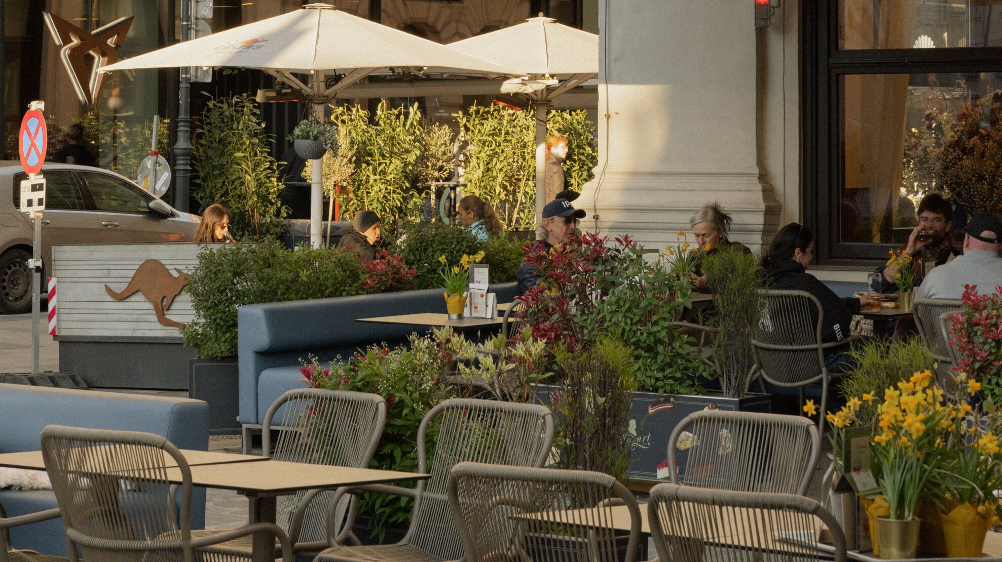 Außenbereich eines Cafés mit Sonnenschirmen und Sitzgelegenheiten, Menschen genießen bei warmem Licht Speisen und Getränke in entspannter Atmosphäre.