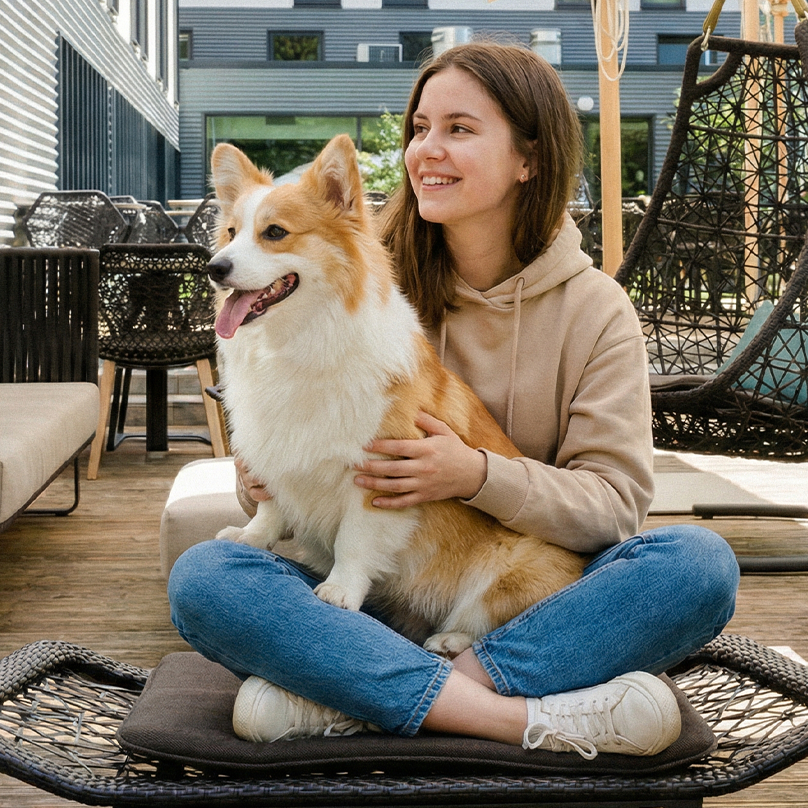 Junge Frau sitzt entspannt auf Terrasse unter Sonnenschirmen, hält glücklich einen Hund auf dem Schoß. Moderne Außenmöbel und freundliche Atmosphäre laden zum Verweilen ein.
