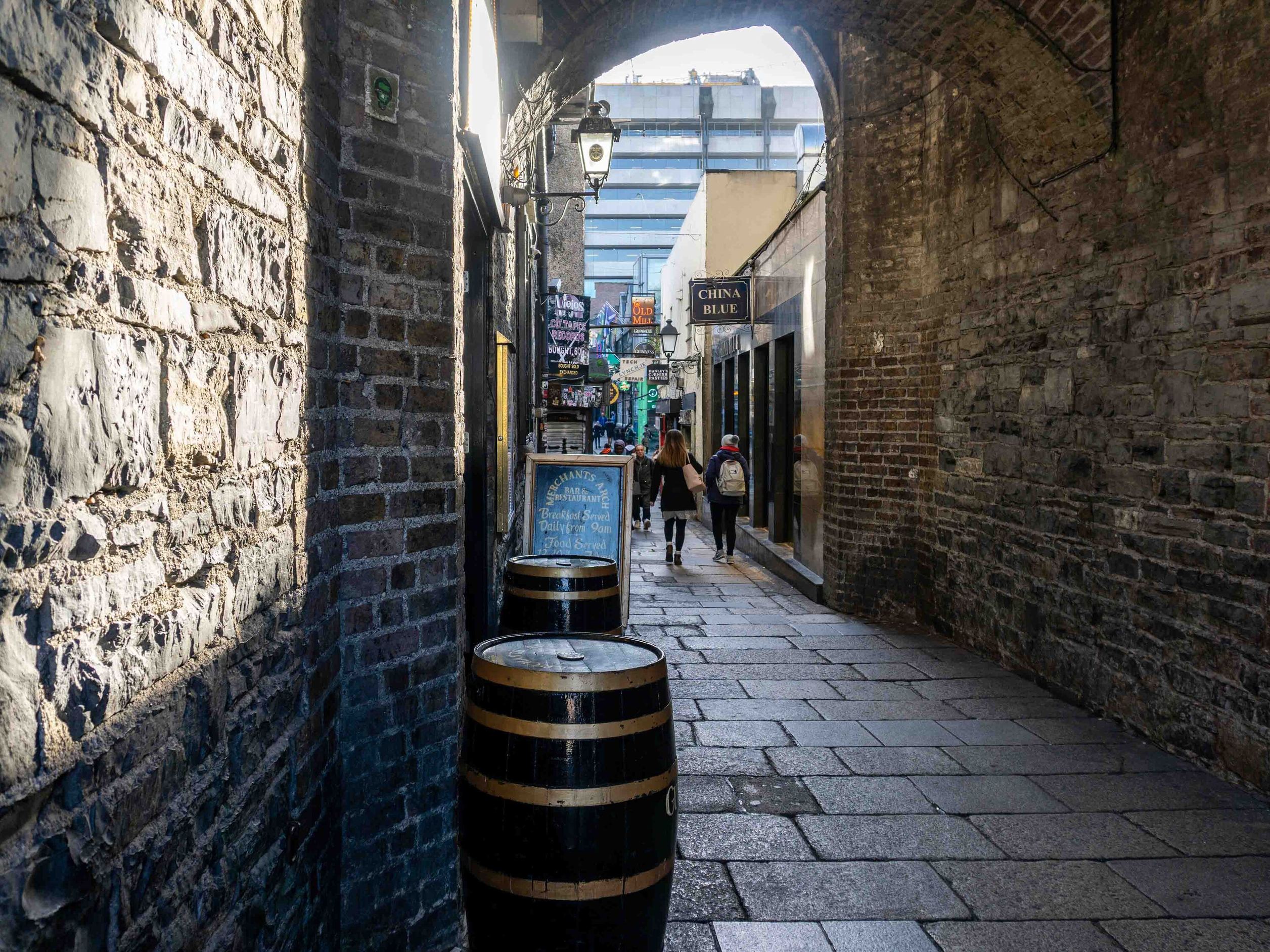 Enge, gepflasterte Gasse mit historischen Steinmauern und Fassfässern, die eine gemütliche, atmosphärische Altstadtkulisse vermitteln, sichtbar bei Tageslicht mit wenigen Passanten.
