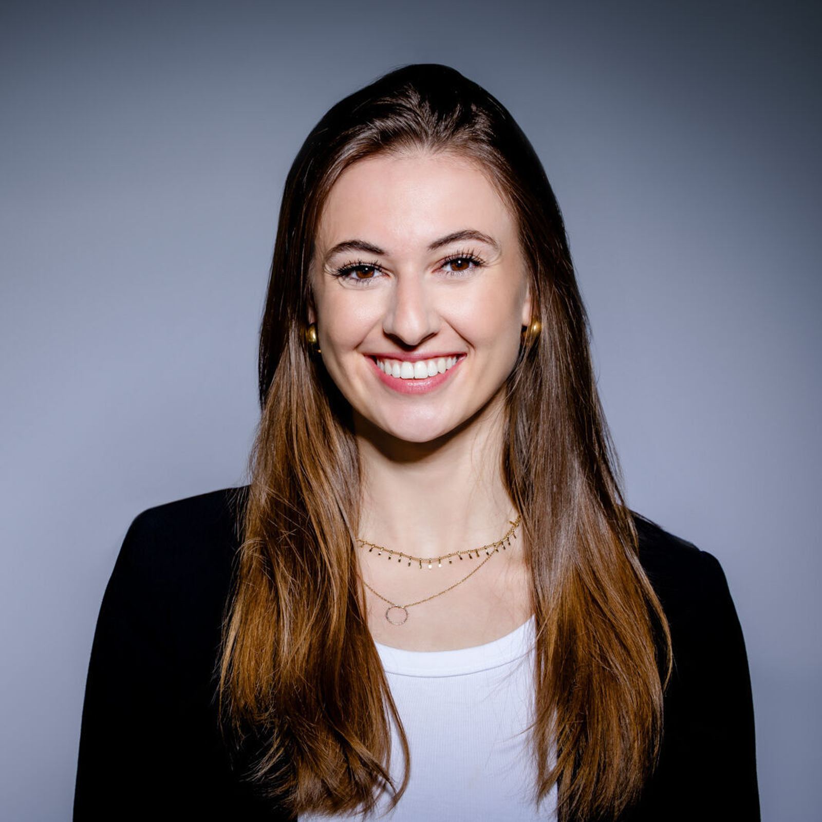 Porträt einer jungen Frau mit langen braunen Haaren, die freundlich lächelt, vor neutralem Hintergrund, trägt ein weißes Oberteil und eine schwarze Jacke.