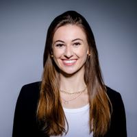 Porträt einer jungen Frau mit langen braunen Haaren, die freundlich lächelt, vor neutralem Hintergrund, trägt ein weißes Oberteil und eine schwarze Jacke.