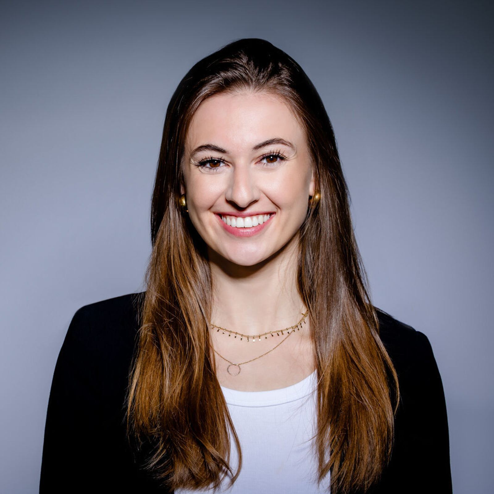 Porträt einer jungen Frau mit langen braunen Haaren, die freundlich lächelt, vor neutralem Hintergrund, trägt ein weißes Oberteil und eine schwarze Jacke.