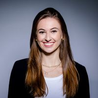 Porträt einer jungen Frau mit langen braunen Haaren, die freundlich lächelt, vor neutralem Hintergrund, trägt ein weißes Oberteil und eine schwarze Jacke.