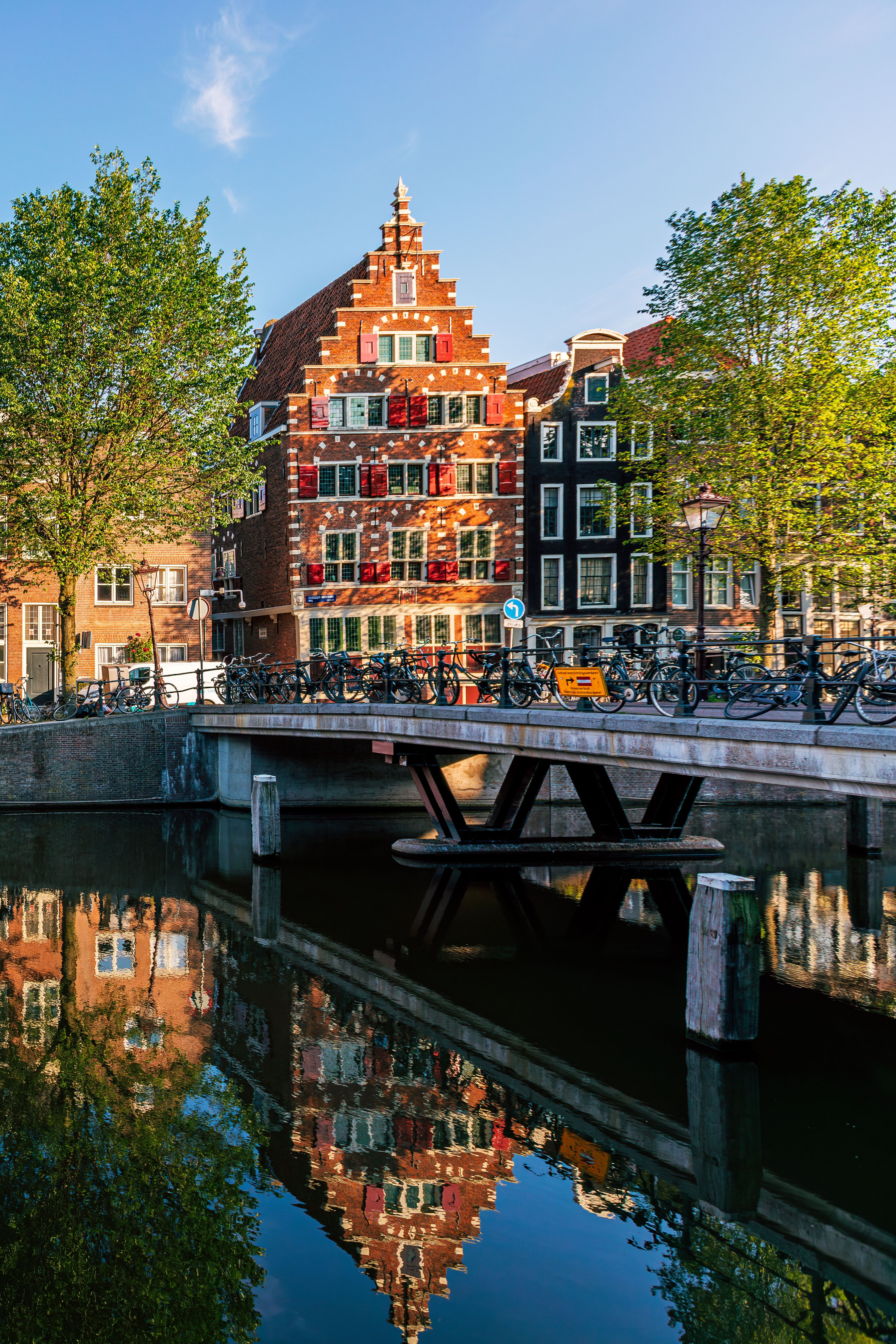 Typische schmale, historische Häuser am Ufer eines ruhigen Kanals in Amsterdam, spiegeln sich im klaren Wasser, begleitet von grün belaubten Bäumen und einer kleinen Brücke bei sonnigem Wetter.