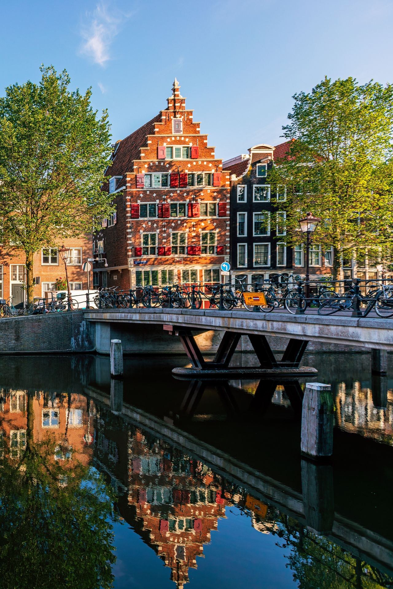 Typische schmale, historische Häuser am Ufer eines ruhigen Kanals in Amsterdam, spiegeln sich im klaren Wasser, begleitet von grün belaubten Bäumen und einer kleinen Brücke bei sonnigem Wetter.