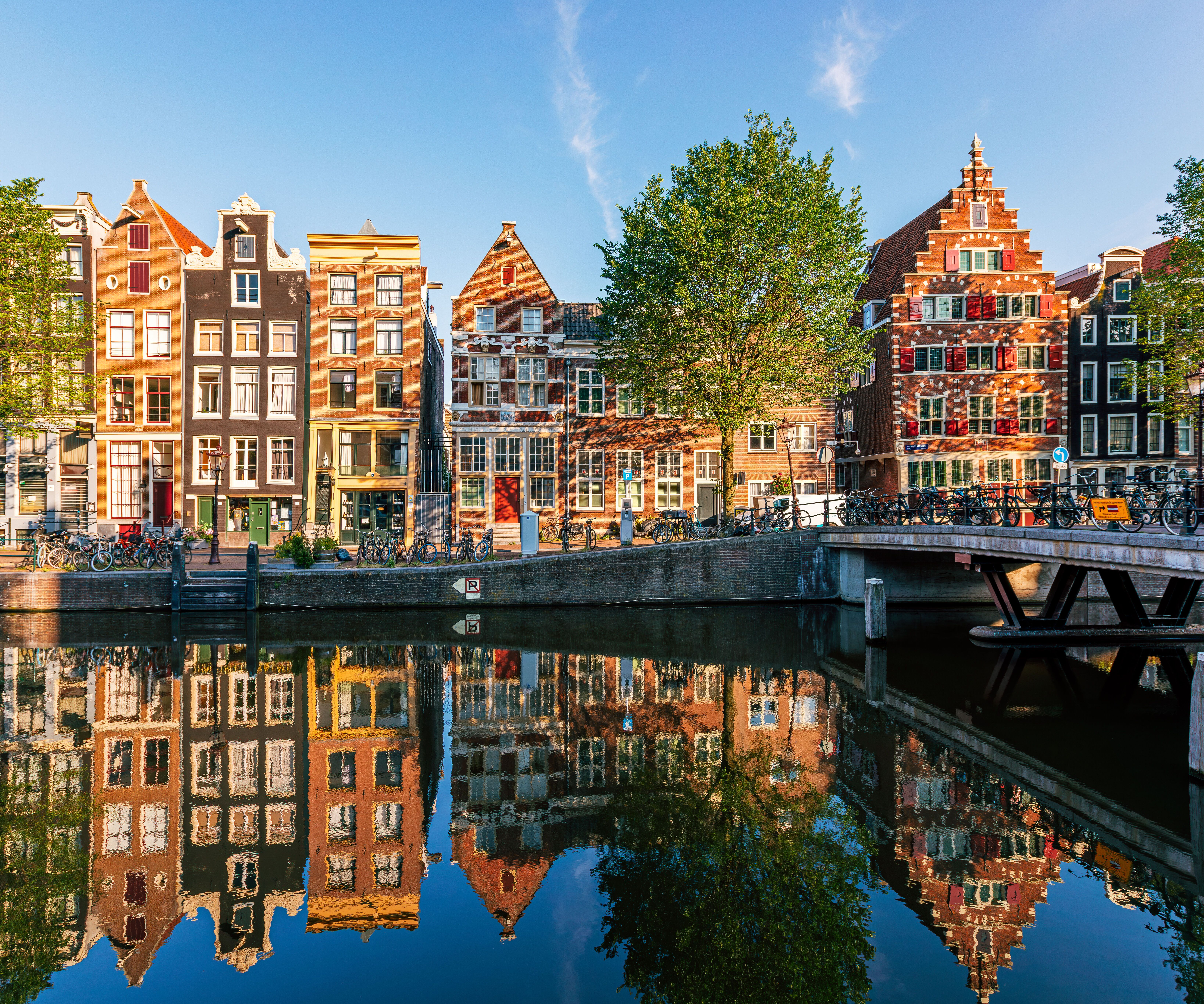 Typische schmale, historische Häuser am Ufer eines ruhigen Kanals in Amsterdam, spiegeln sich im klaren Wasser, begleitet von grün belaubten Bäumen und einer kleinen Brücke bei sonnigem Wetter.