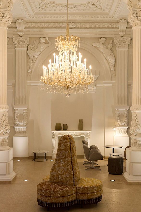 Prächtiger, historisch gestalteter Innenraum mit hoher Stuckdecke, verzierten Säulen und elegantem Kronleuchter. Gemütliche Sitzmöbel laden zum Entspannen ein. Atmosphäre verbindet klassische Pracht mit modernem Komfort.