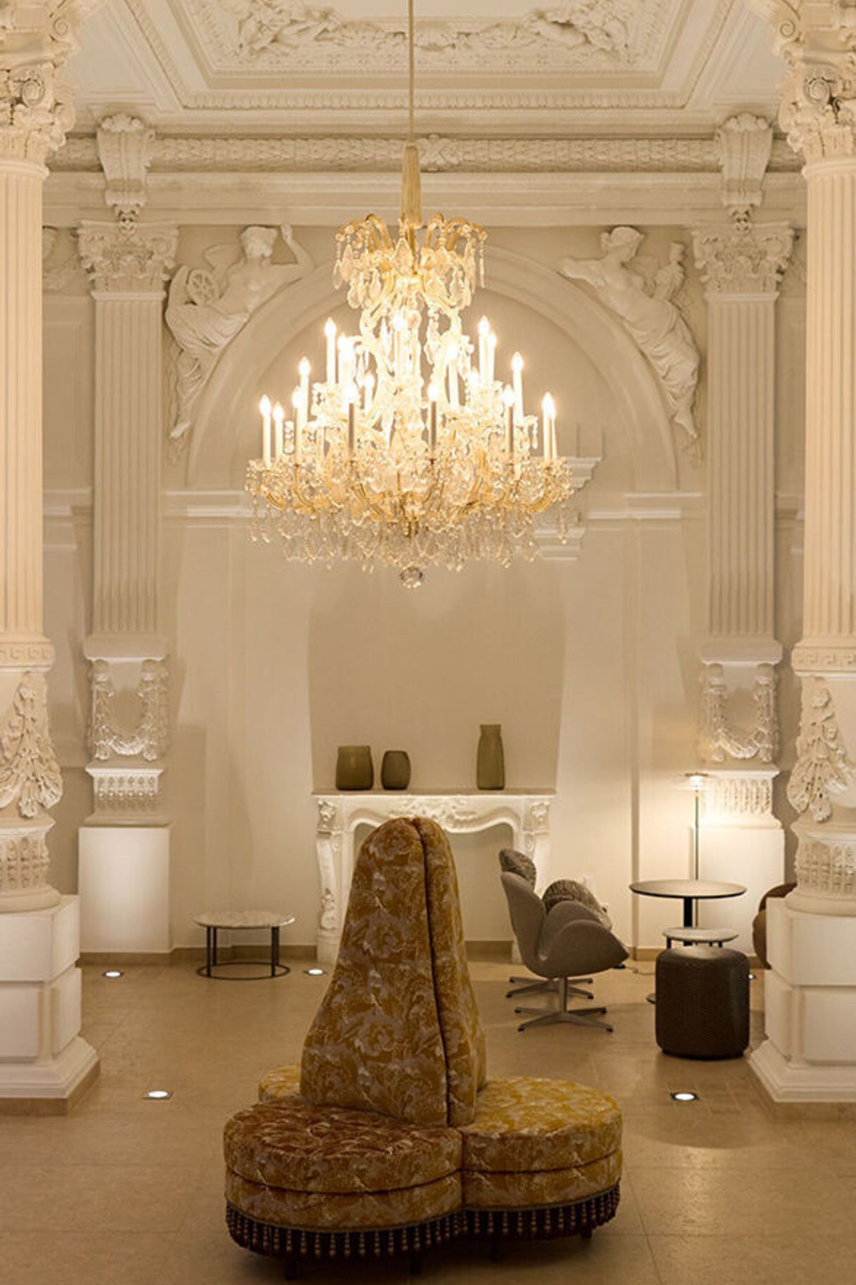 Prächtiger, historisch gestalteter Innenraum mit hoher Stuckdecke, verzierten Säulen und elegantem Kronleuchter. Gemütliche Sitzmöbel laden zum Entspannen ein. Atmosphäre verbindet klassische Pracht mit modernem Komfort.