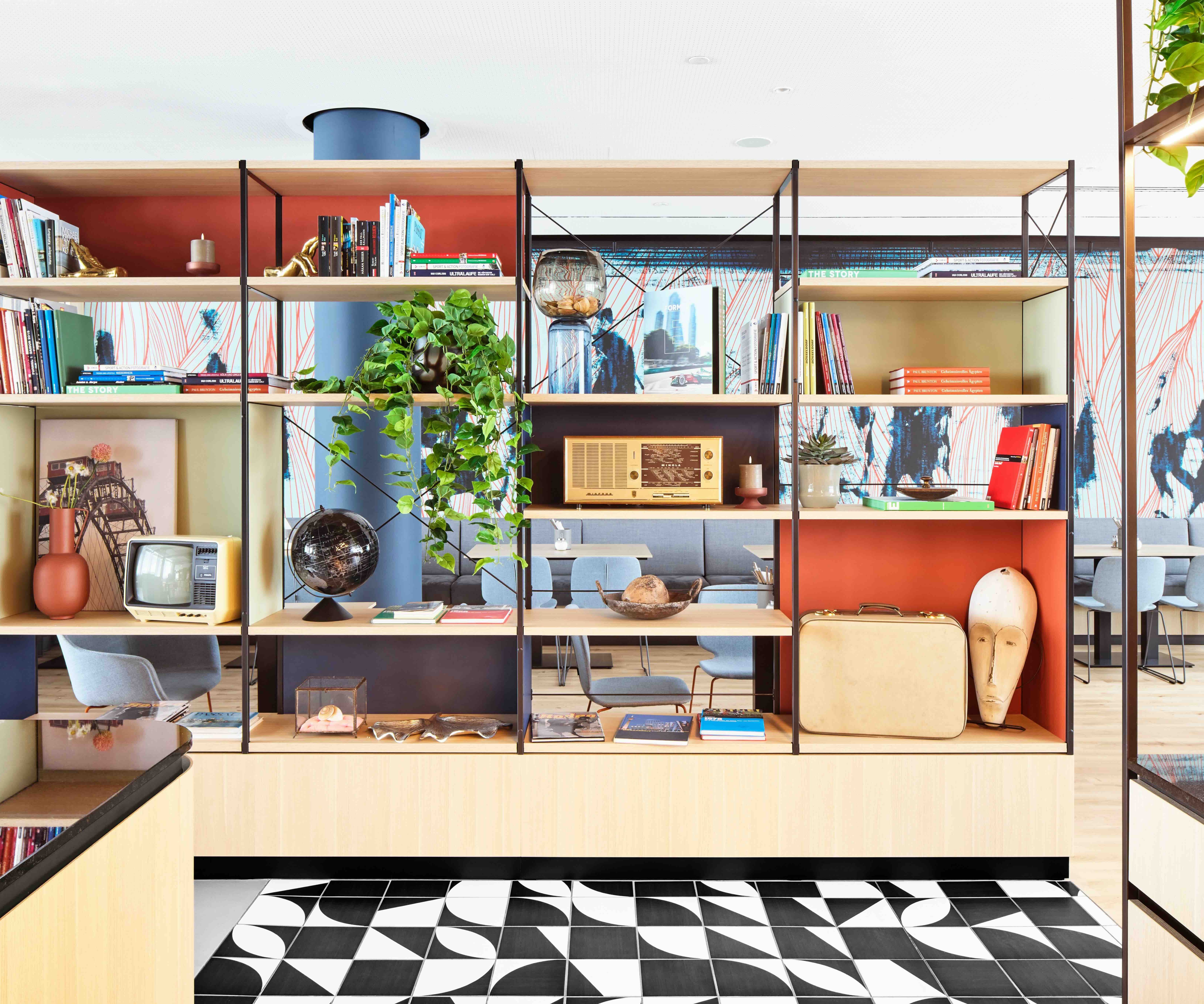 Offene Regalwand mit Büchern, Pflanzen, historischen Radios und Dekorationsobjekten, die eine moderne, gemütliche Atmosphäre im Hotel-Loungebereich schafft.