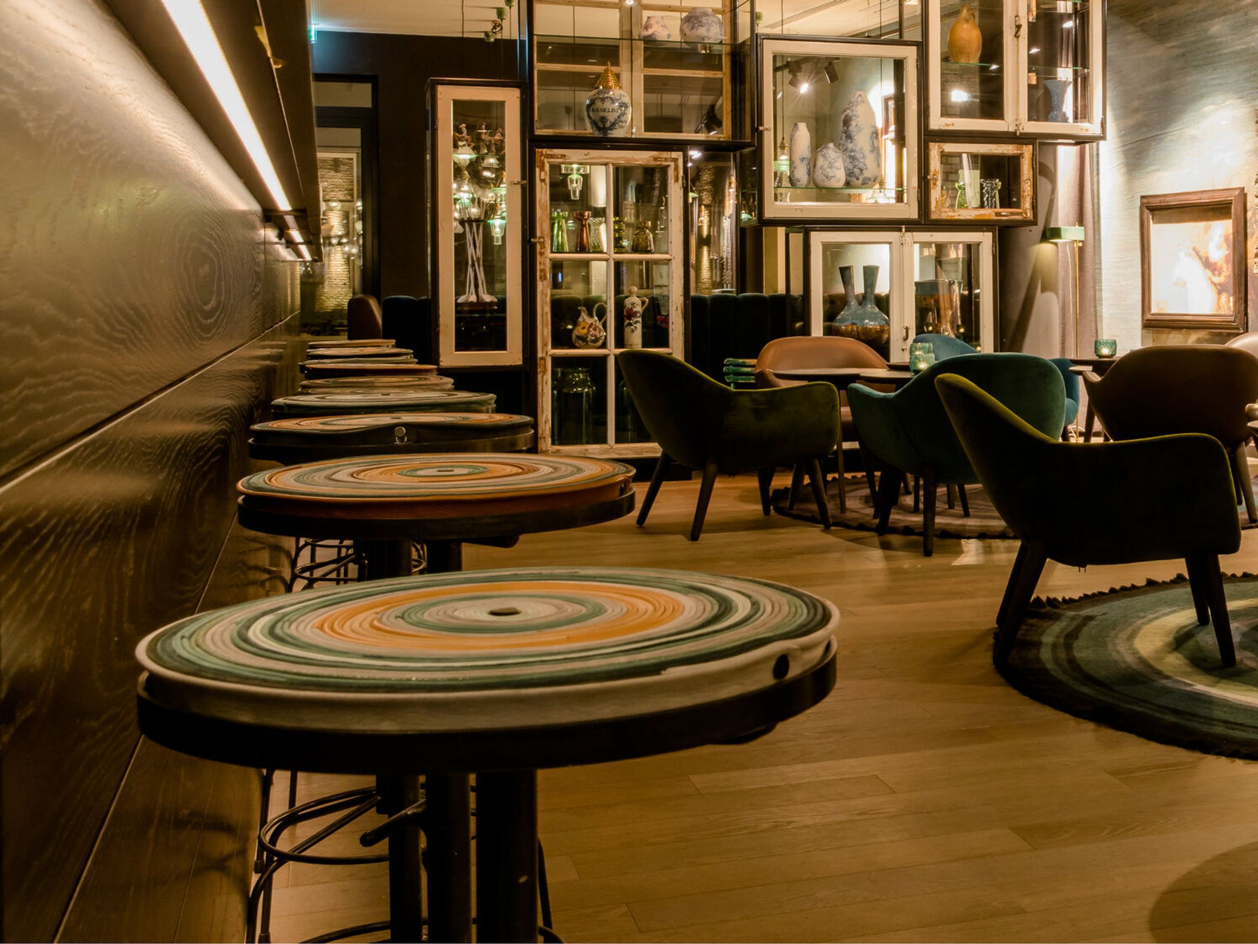 Motel One Hotelbarbereich mit Rundtischen, bequemen Sesseln und warmem Licht, umgeben von dekorativen Vitrinen.