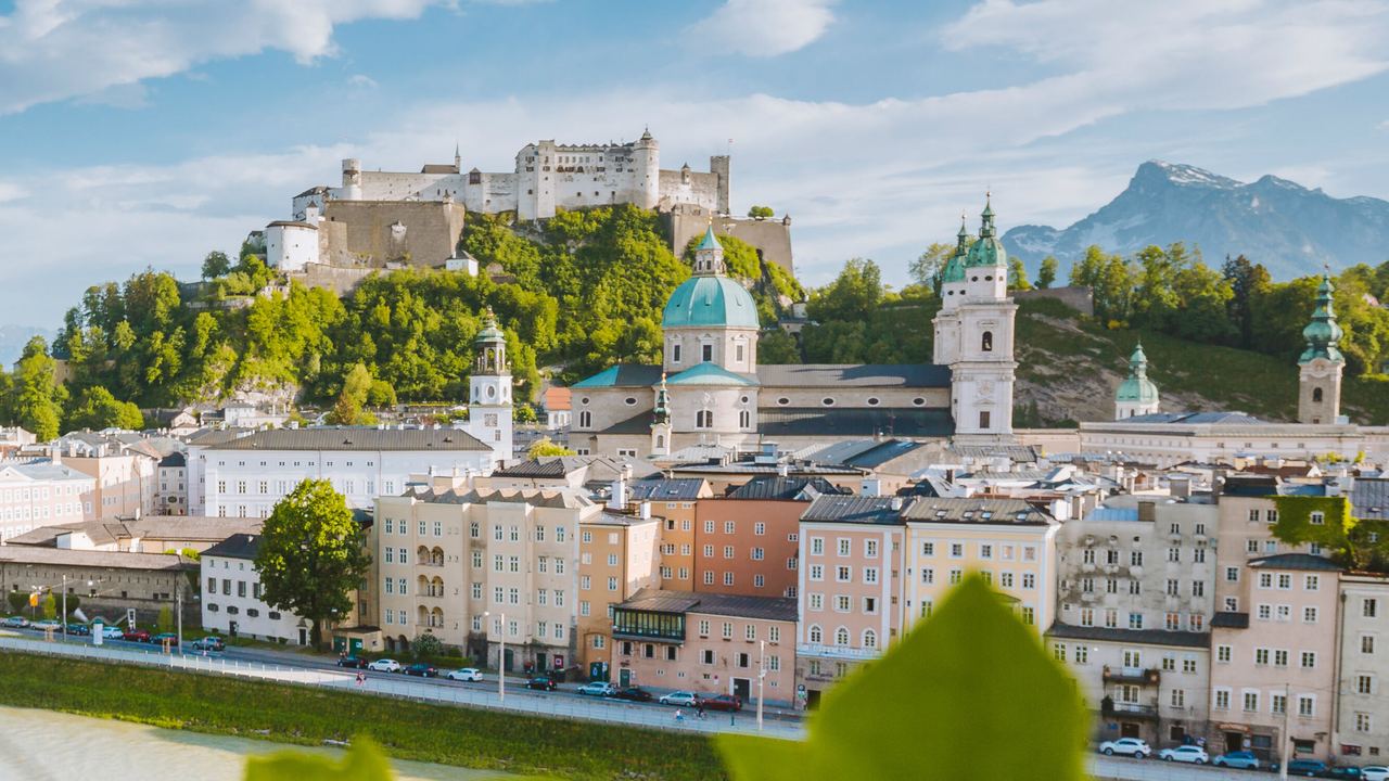 Blick auf die historische Altstadt von Salzburg mit der eindrucksvollen Festung Hohensalzburg auf einem bewaldeten Hügel und den Alpen im Hintergrund, bei klarem Himmel. Im Vordergrund grüne Blätter, die eine natürliche Atmosphäre schaffen.