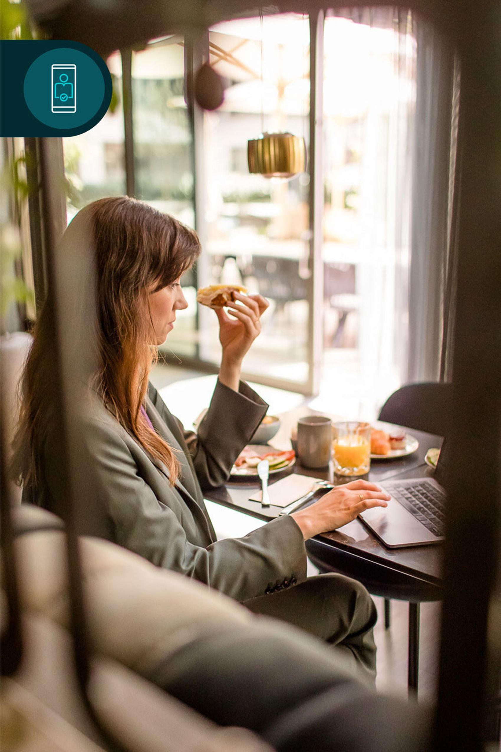 Eine Frau genießt ein Croissant beim Arbeiten an ihrem Laptop in der hellen One Lounge mit großem Fenster. Auf dem Tisch stehen ein Glas Orangensaft und eine Tasse Kaffee.